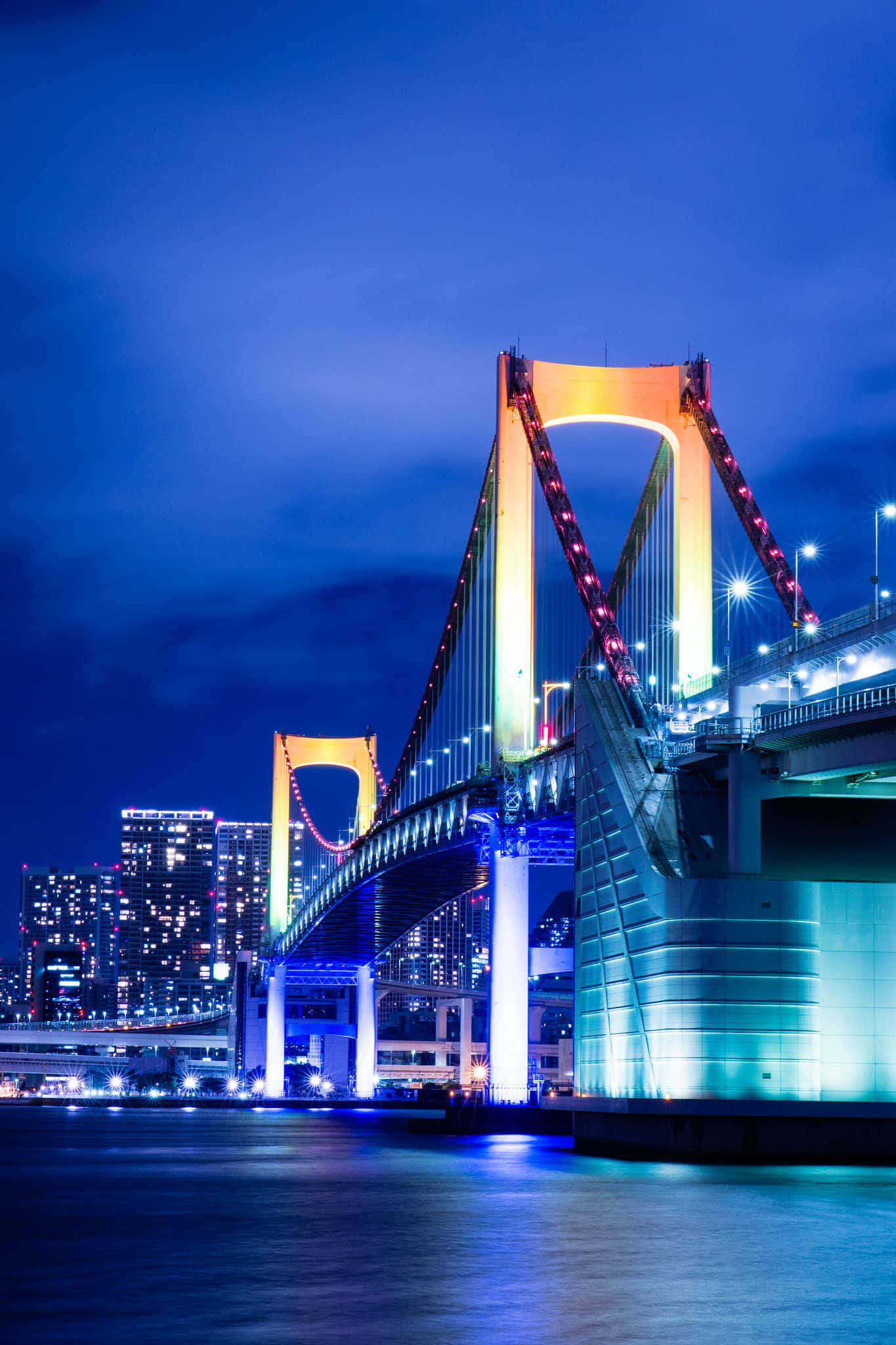 美國猶他州湖粉的壯麗彩虹橋 壁紙 — 東京的彩虹橋在深藍色夜空下閃耀著鮮豔燈光，倒映在平靜的水面上。
