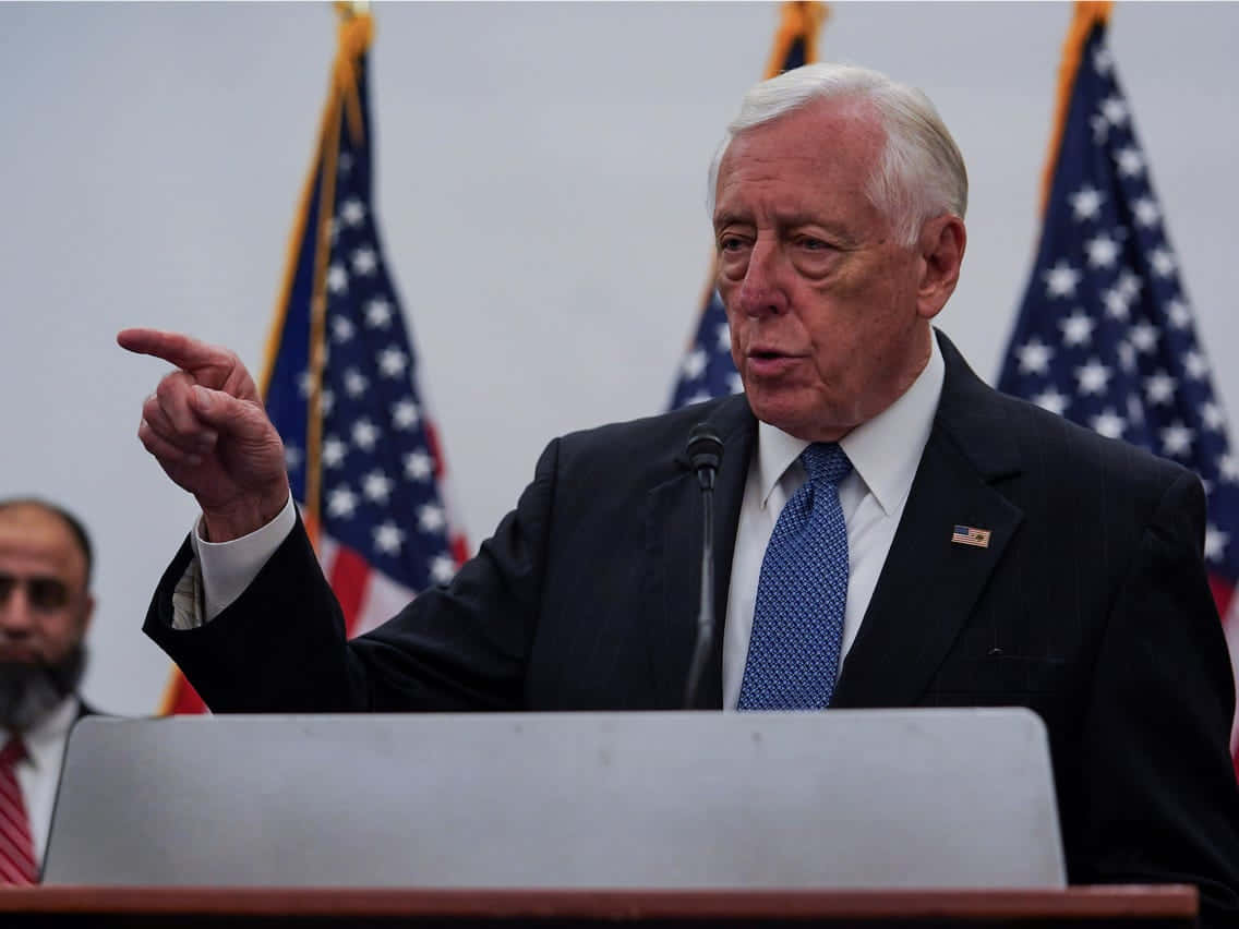 美國眾議員 Steny Hoyer 熱情演說 壁紙 — 一位身穿深色西裝和藍色領帶的年長男性，在身後有美國國旗的講台上，以強調的手勢發表演說。