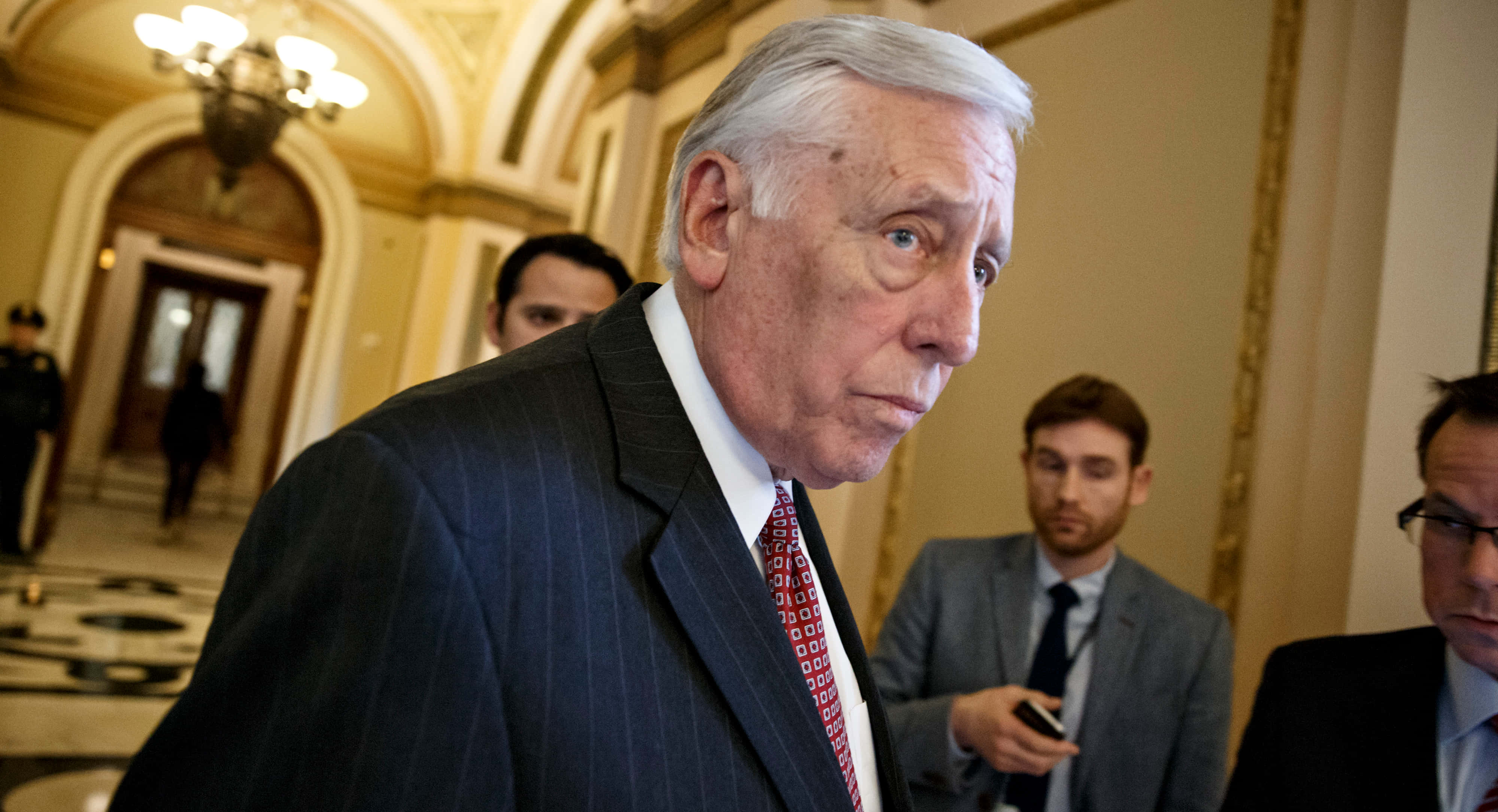 美國眾議院 Majority Leader 斯泰尼·霍耶側影 壁紙 — 一位身穿深色細條紋西裝和紅色圖案領帶的年長男士，走過一條裝飾華麗的走廊，走廊有拱形門口和吊燈。