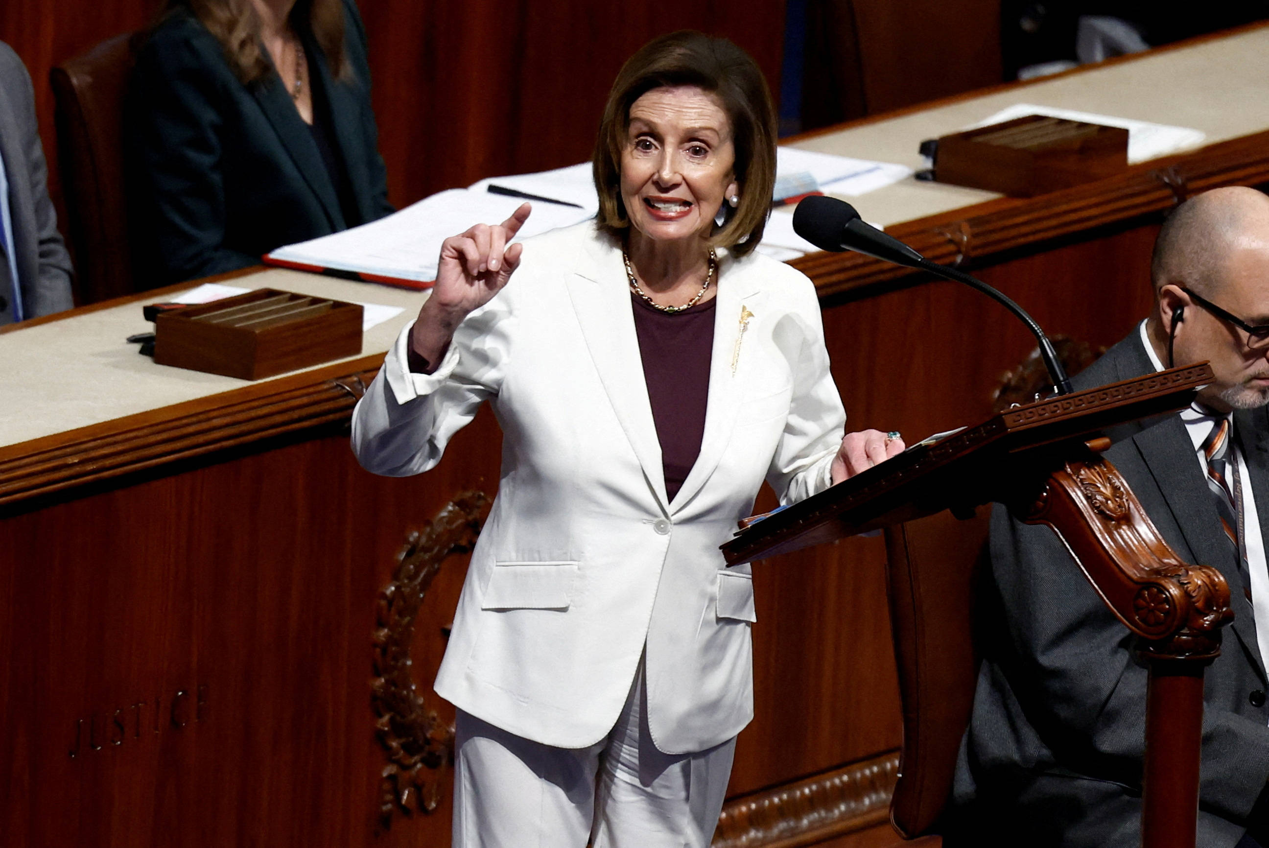 美國眾議院議長南希·佩洛西穿時尚白色西裝褲 壁紙 — 蘭茜·佩洛西在一個裝飾華麗木工、正式的立法會議廳內，於木製講台上充滿激情地發言，附近有專注的工作人員。
