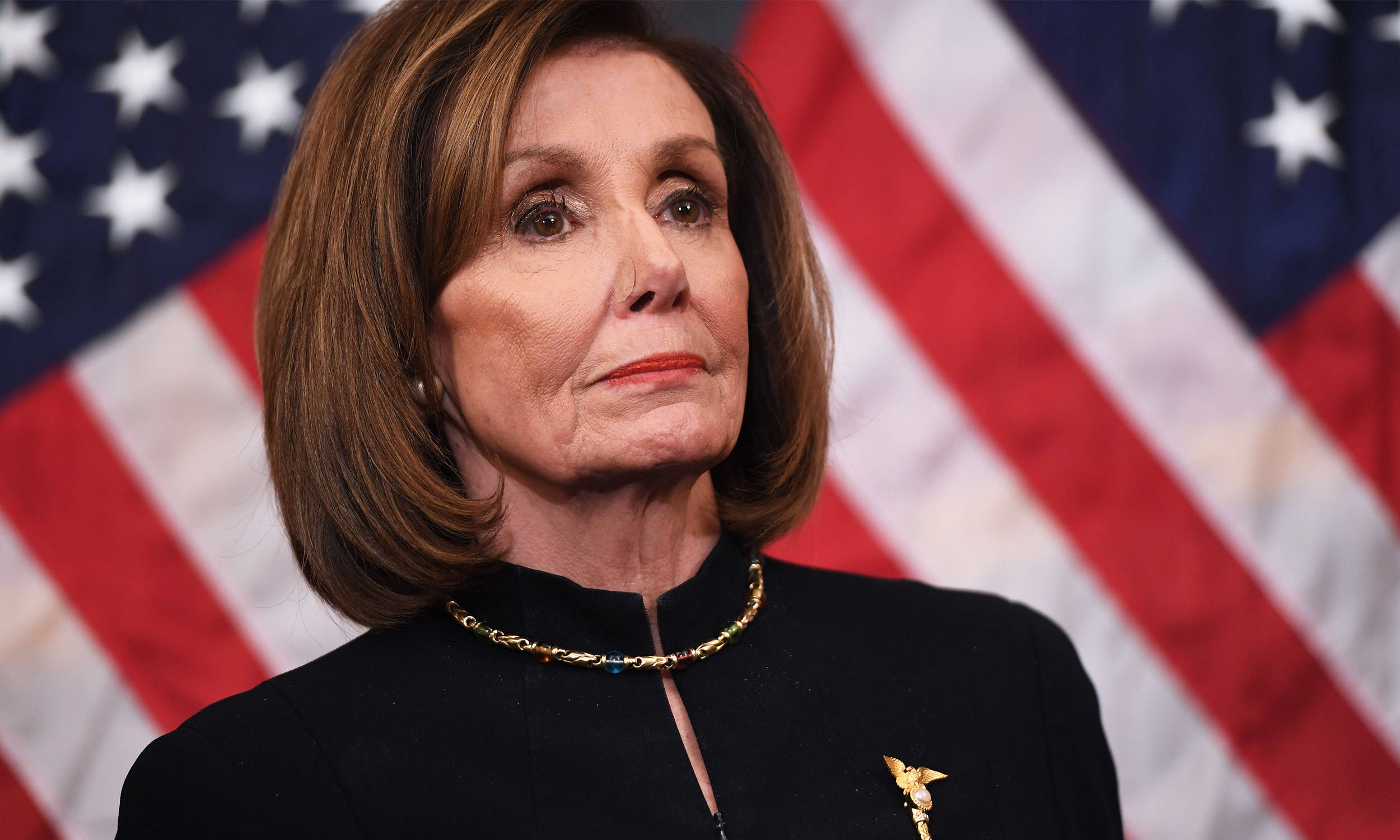 美國眾議院議長南希·佩洛西身穿黑色禮服立於美國國旗前 壁紙 — 蘭茜·佩洛西身穿黑色西裝和金色項鍊，莊嚴地站在美國國旗前。