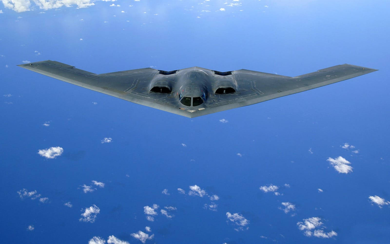 美國空軍 B-2 壁紙 — 一架流線型灰色隱形轟炸機在廣闊的藍天中翱翔，點綴著散落的白色雲朵。