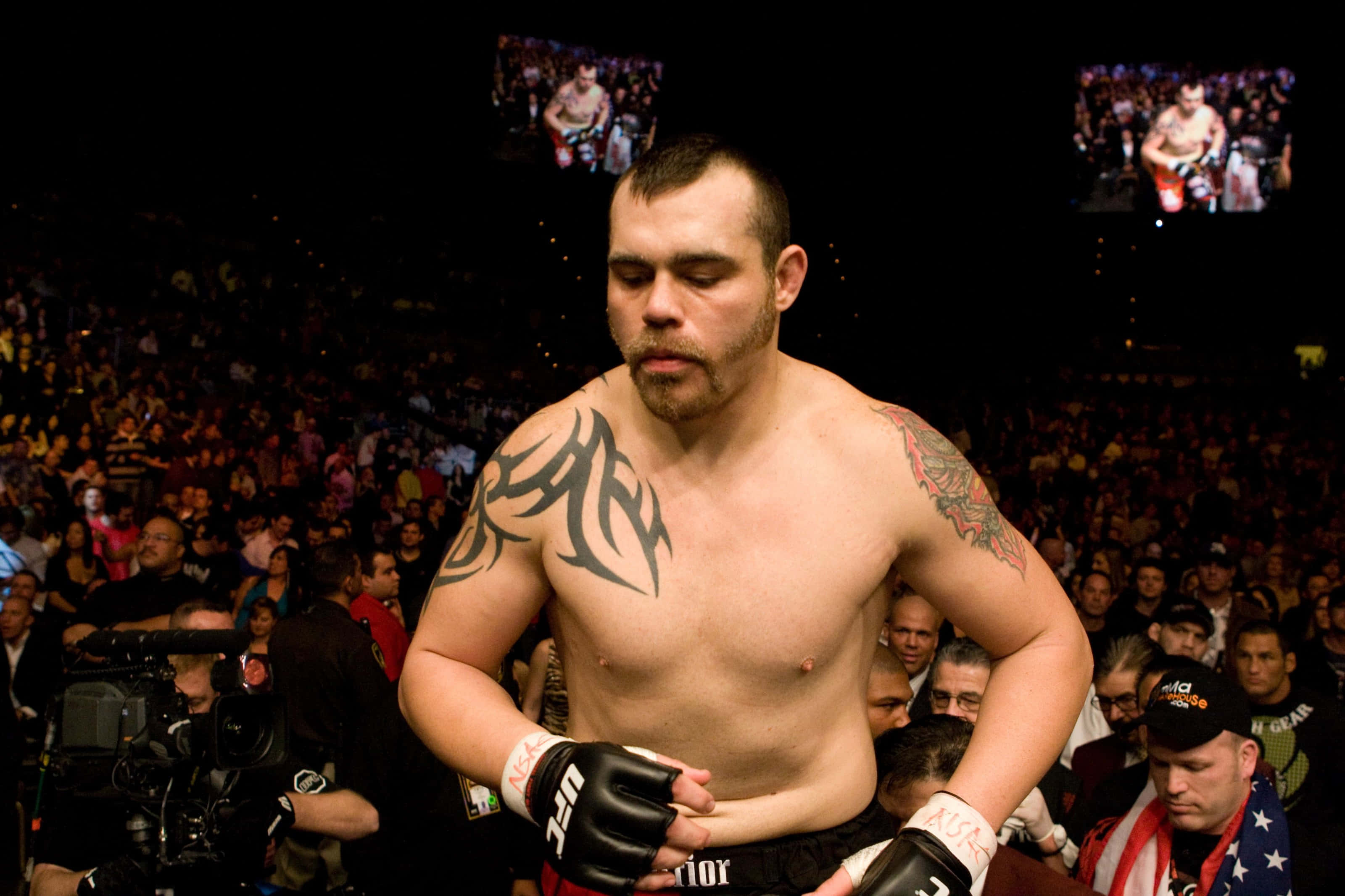 美國綜合格鬥選手 Tim Sylvia 踏入八角籠 壁紙 — 一位紋身的MMA拳手戴著黑色手套，在場館燈光下專注地站在咆哮的人群前。