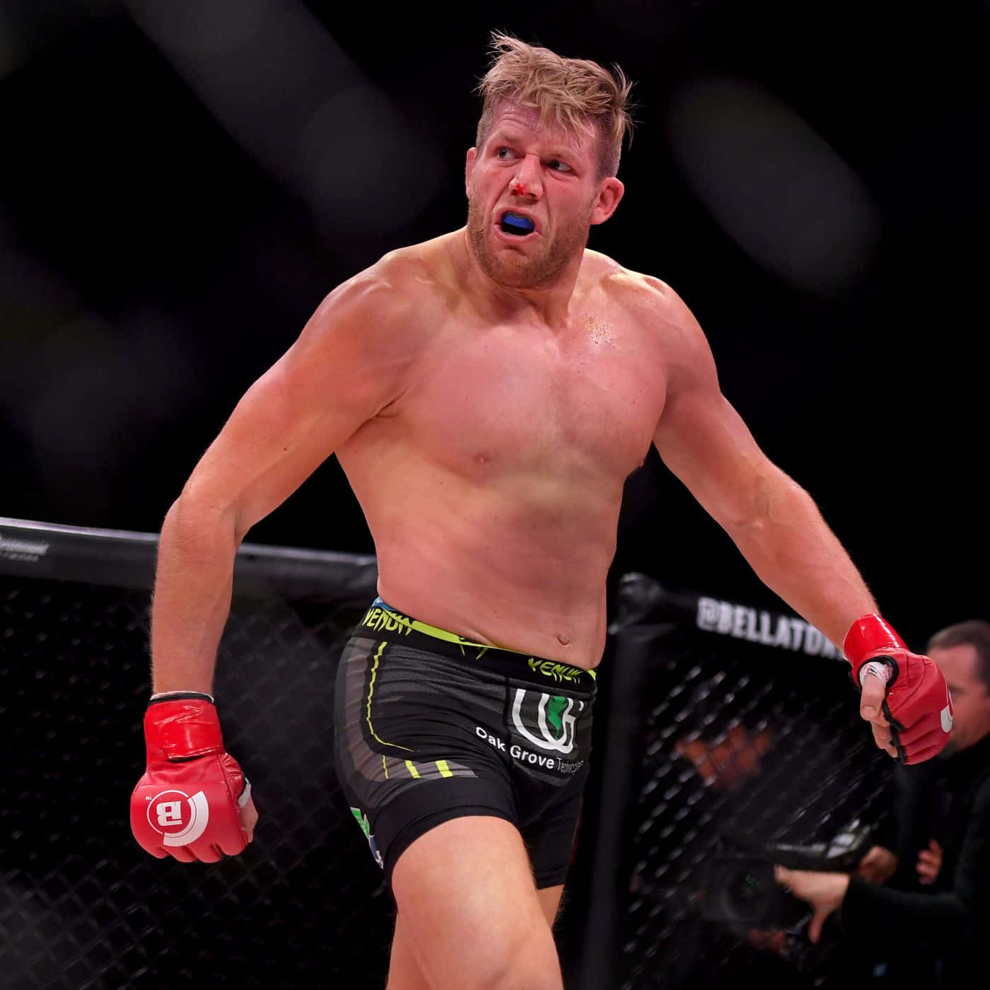 美國綜合格鬥選手傑克·哈格於Bellator擂台內 壁紙 — 一名肌肉發達的戰士戴著藍色護齒套，在燈光昏暗的八角籠中咆哮勝利。