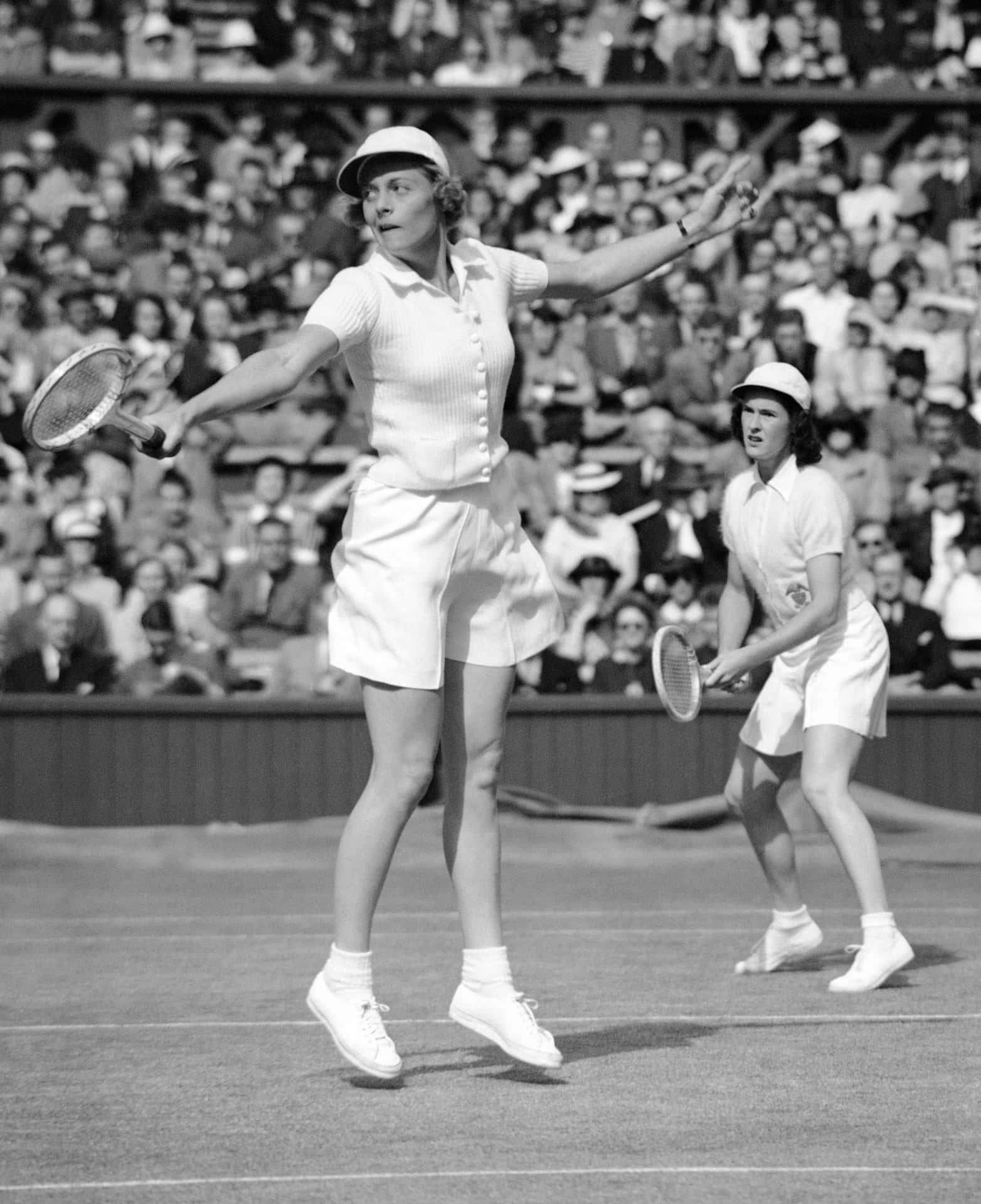 美國網球選手愛麗絲·馬伯爾於1939年溫布爾登 壁紙 — 兩位身穿復古白色網球服裝的女性在草地上進行雙打比賽，周圍擠滿了觀眾。