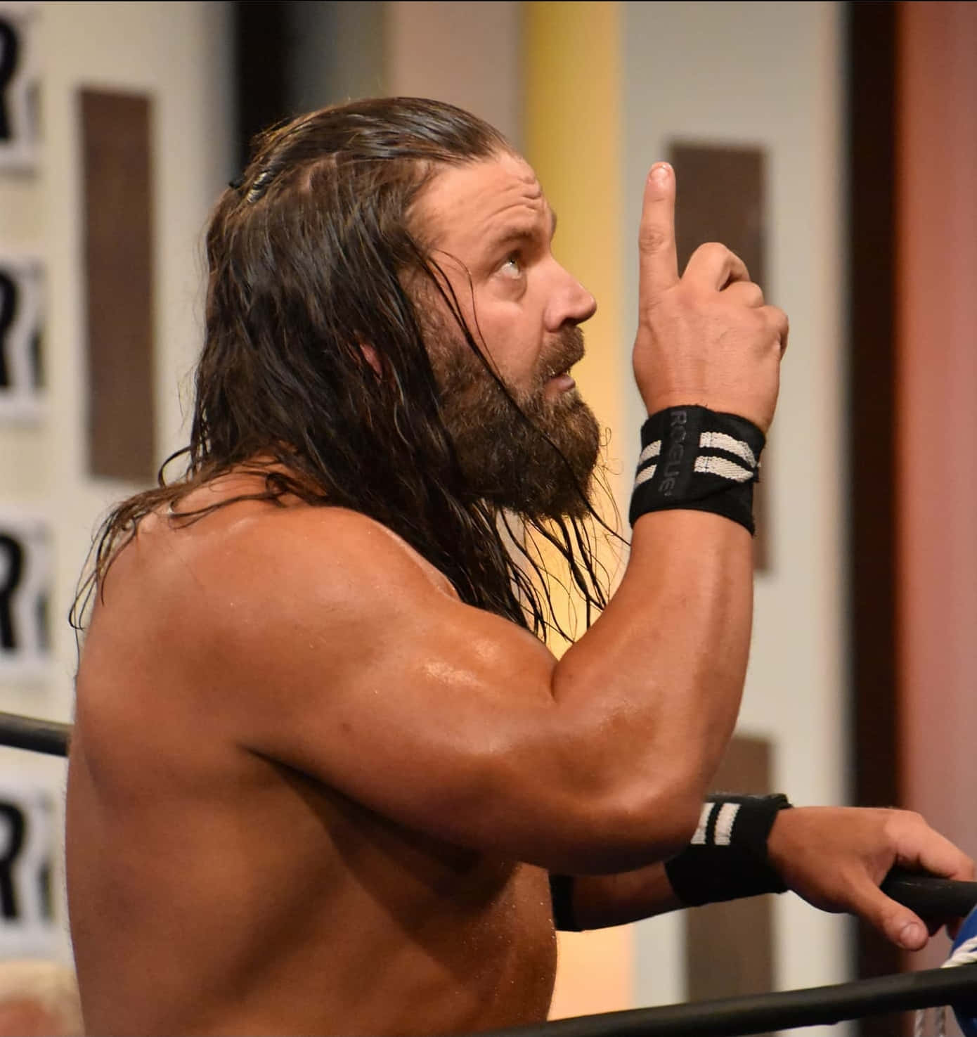 美國職業摔角手James Storm於私人健身房 壁紙 — 一位肌肉發達、留鬍鬚的摔角手，濕髮向上指，同時抓住光線充足的競技場中的擂台繩索。