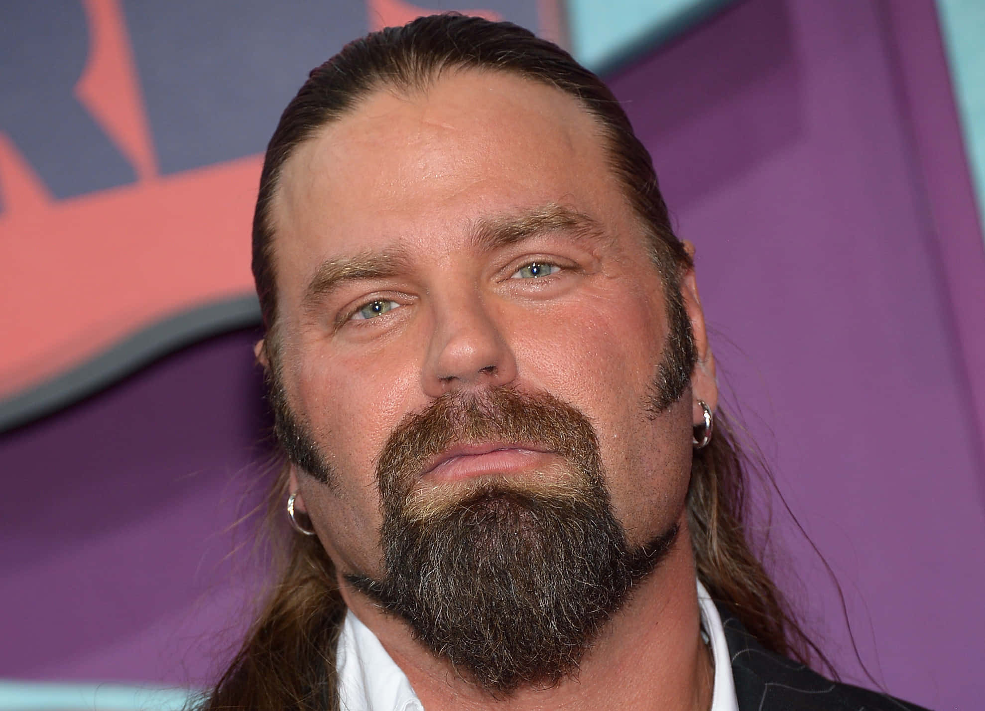 美國職業摔角手James Storm出席2014 CMT音樂獎 壁紙 — 一名留著深色長髮和滿臉鬍鬚、戴著耳環的男子，在紫色和橙色背景前的特寫肖像。