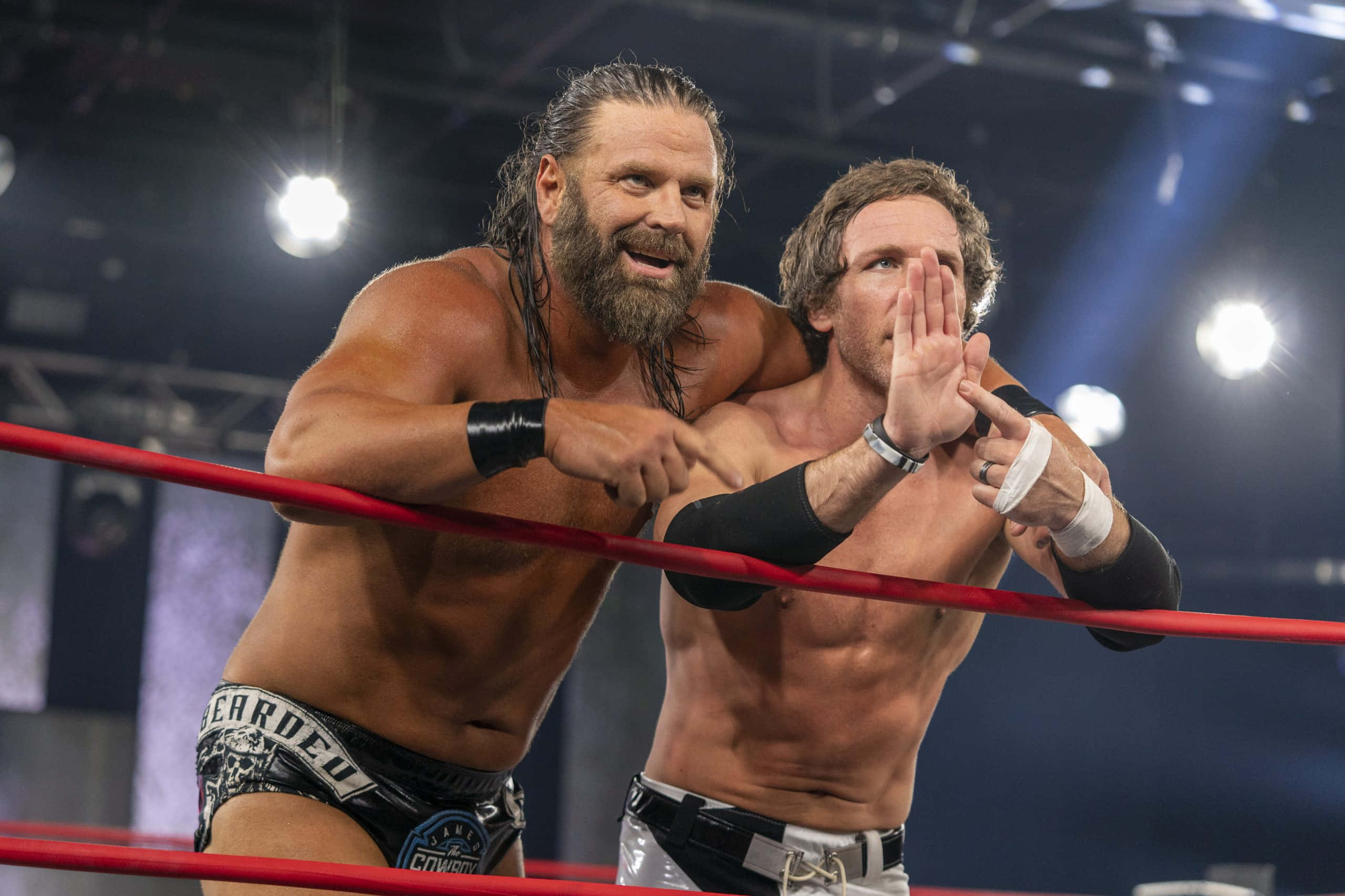美國職業摔角手James Storm與Chris Sabin賽前合影 壁紙 — 兩名肌肉發達的摔角手在擂台上，其中一人指責地指向對方，另一人則舉手抗議，在明亮的場館燈光下。