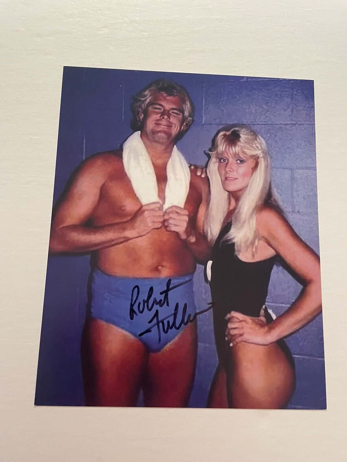 美國職業摔角手Robert Fuller與Miss Sylvia簽名照 壁紙 — 一張摔角手 Robert Fuller 與一位金髮女子在藍色煤渣磚牆前擺姿勢的簽名照片。