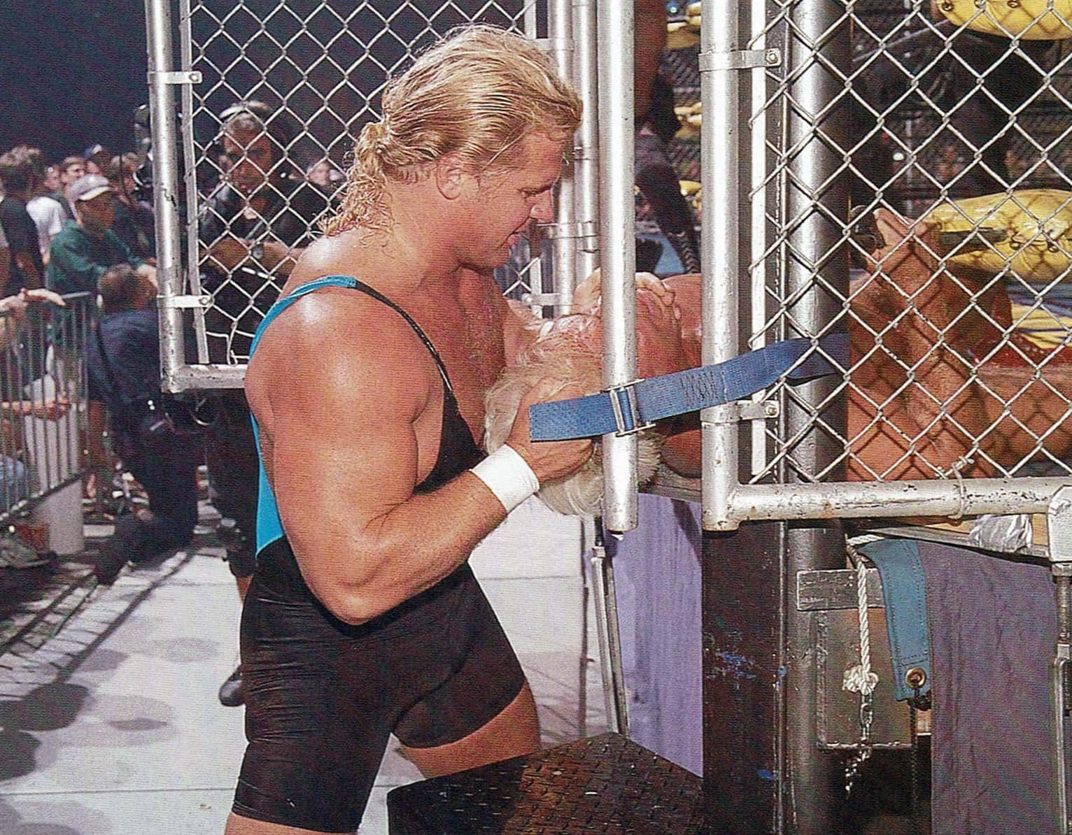 美國職業摔角手庫特·亨尼格 WCW WarGames 壁紙 — 一位肌肉發達的金髮摔角手穿著藍黑相間的緊身衣，在明亮的場館燈光下，於摔角比賽中抓住鐵鏈籠門。
