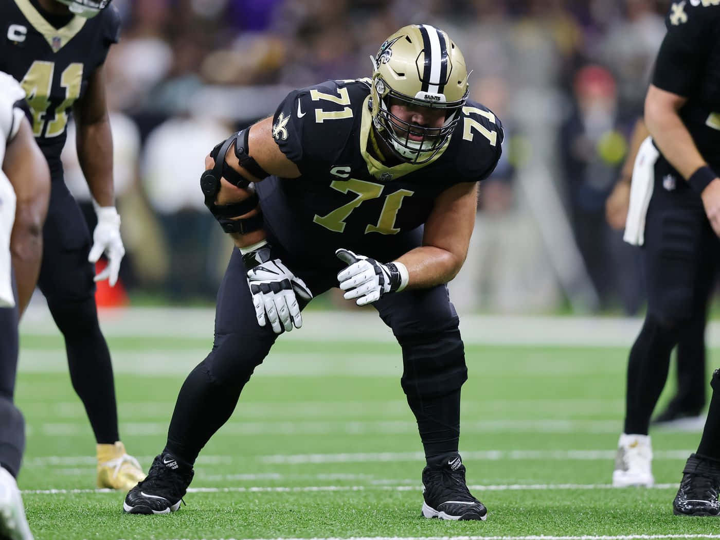 美國職業美式足球員Ryan Ramczyk對戰巴爾的摩烏鴉隊 壁紙 — 新奧爾良聖徒隊的進攻鋒線球員身穿黑色和金色裝備，在比賽期間低蹲在綠色球場上。