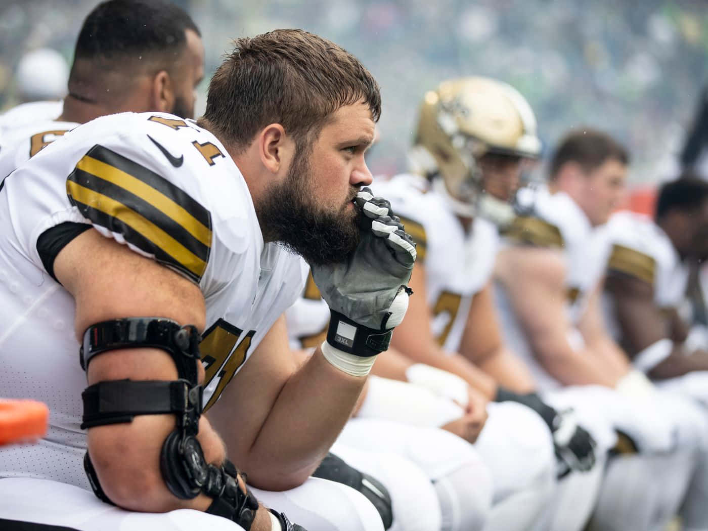 美國職業美式足球員Ryan Ramczyk坐於板凳觀賽 壁紙 — 一位專注的足球運動員身穿白色和金色球衣，在比賽中坐在板凳上，手放在嘴上。