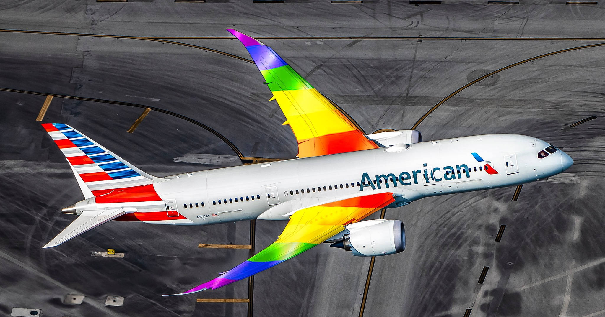 美國航空彩虹翅膀 壁紙 — 一架美國航空公司的飛機帶有彩虹色機翼，在明亮的日光下滑行在灰色的機場停機坪上。