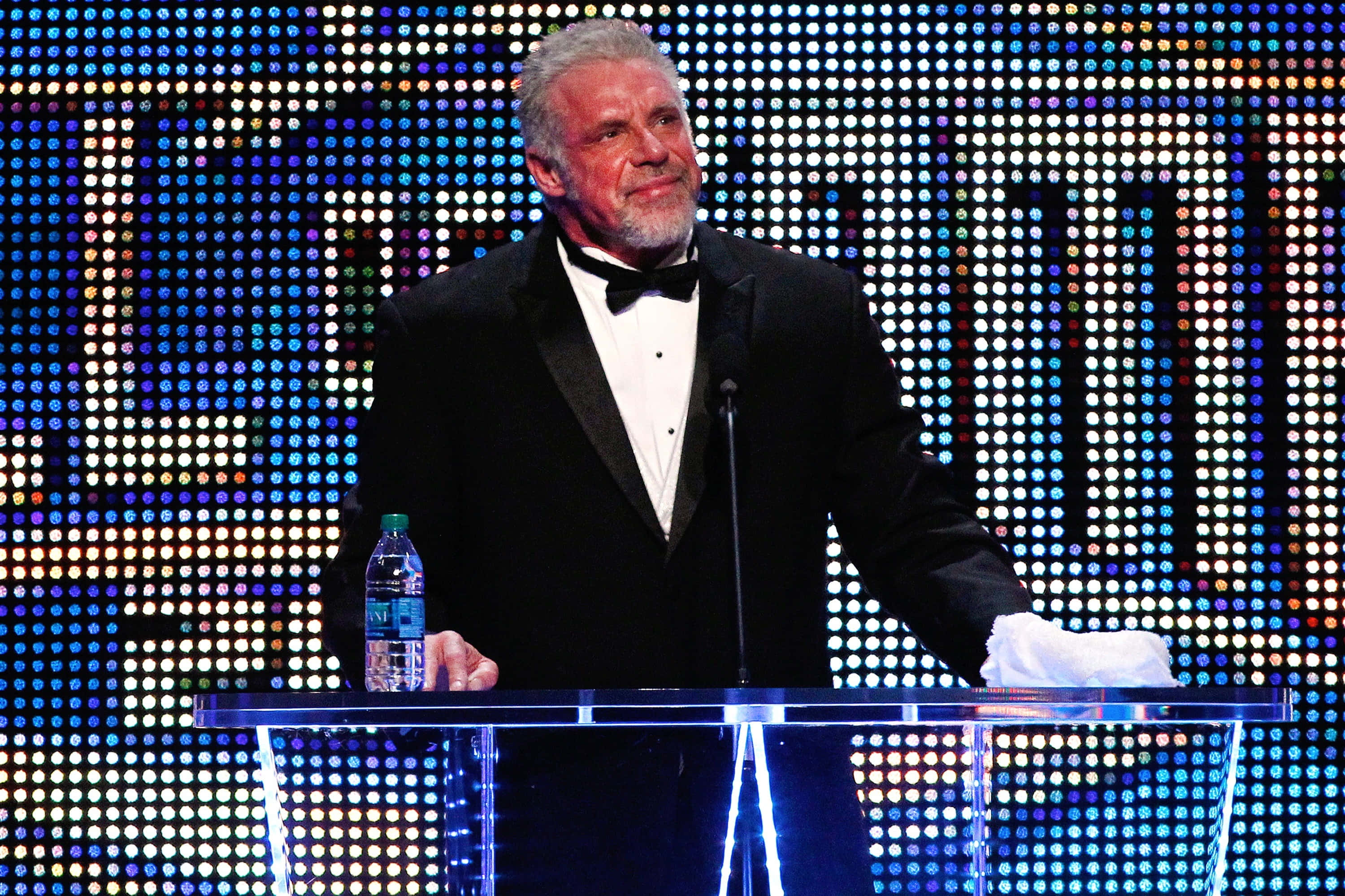 美國資深摔角手 Ultimate Warrior WWE名人堂入選儀式 壁紙 — 一位身穿燕尾服的男士站在透明講台前，背景是耀眼的多彩燈光網格。