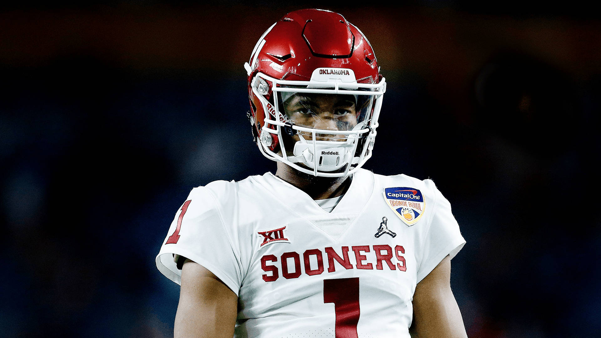 美國足球員凱勒·默里嚴肅表情 壁紙 — 一位專注的奧克拉荷馬大學 Sooners 美式足球員，身穿白色和紅色裝備，在體育館燈光下凝視。