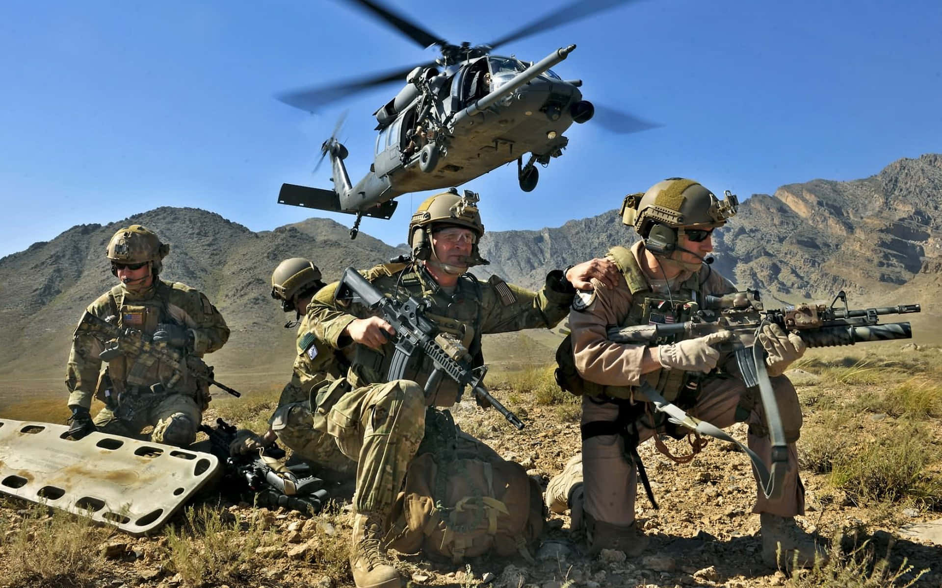 穿著沙漠裝備的士兵蹲下持步槍，一架直升機在岩石地形上空盤旋。
