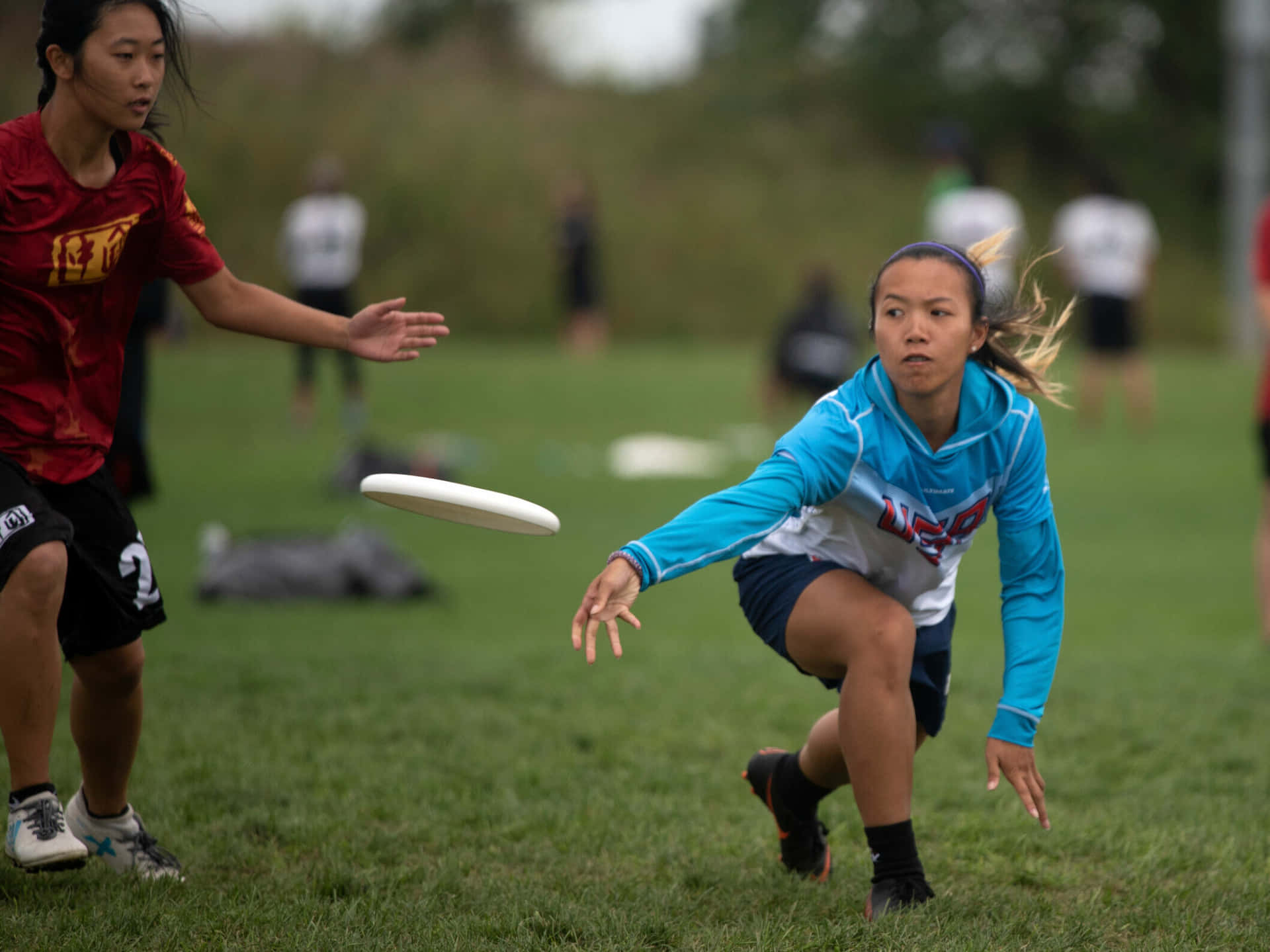美國隊女子 Ultimate Frisbee 運動員 1080p 背景 壁紙 — 兩名女運動員在草地上激烈競爭，一個白色飛盤在戶外比賽中在她們之間飛過。