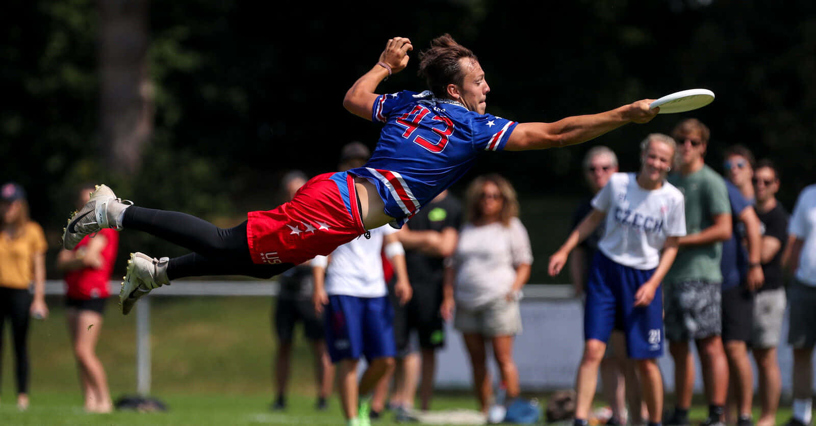 美國隊最佳 Ultimate Frisbee 背景 壁紙 — 一名身穿藍、紅、白三色制服嘅男性終極飛盤選手跳起接住飛盤，背景有觀眾觀看。