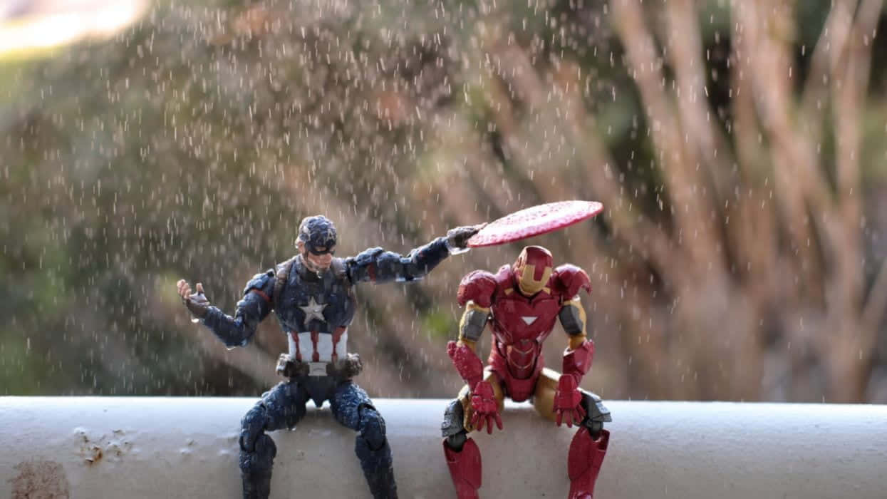 Iron Man Action Figures 桌布 壁紙 — 美國隊長和鐵甲奇俠的動作人偶並排坐在壁架上，雨滴在模糊的自然背景中落在它們周圍。