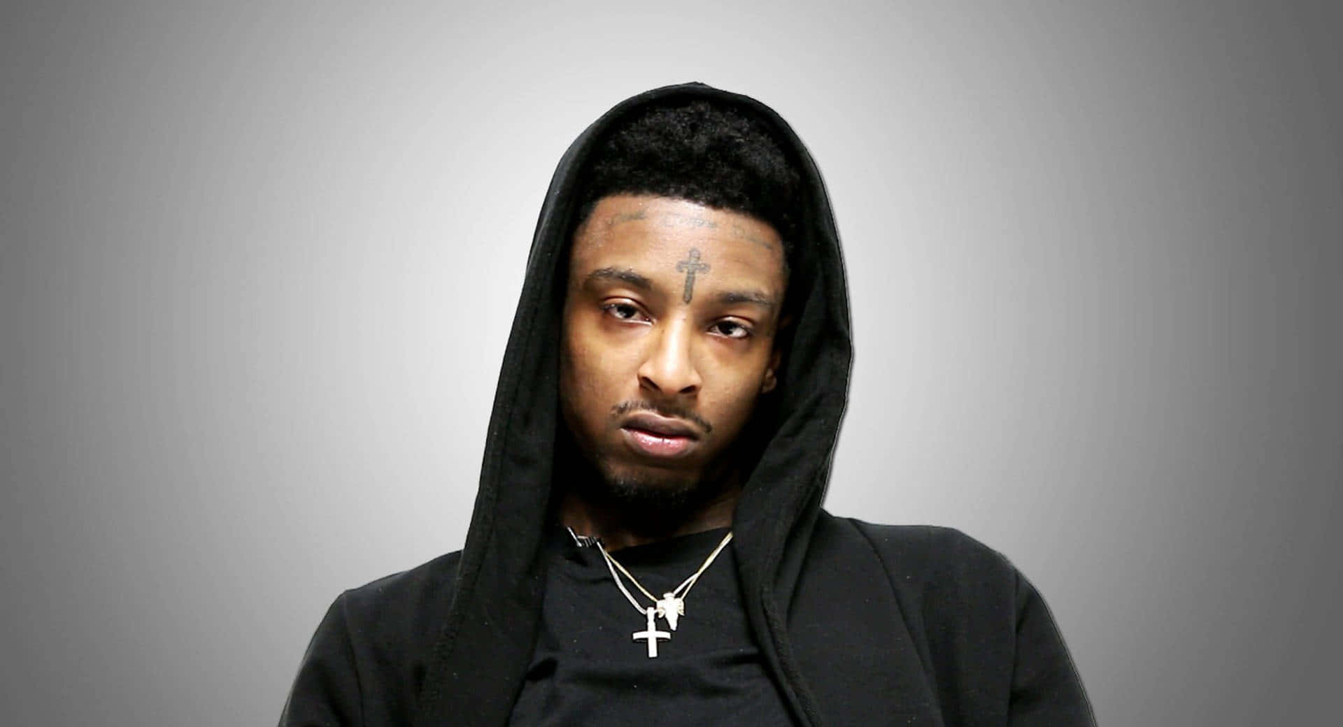 美國饒舌歌手 21 Savage 的形象照 壁紙 — 一個穿著黑色連帽衫、額頭上有十字架紋身的男人，在純灰色背景下強烈地凝視著。