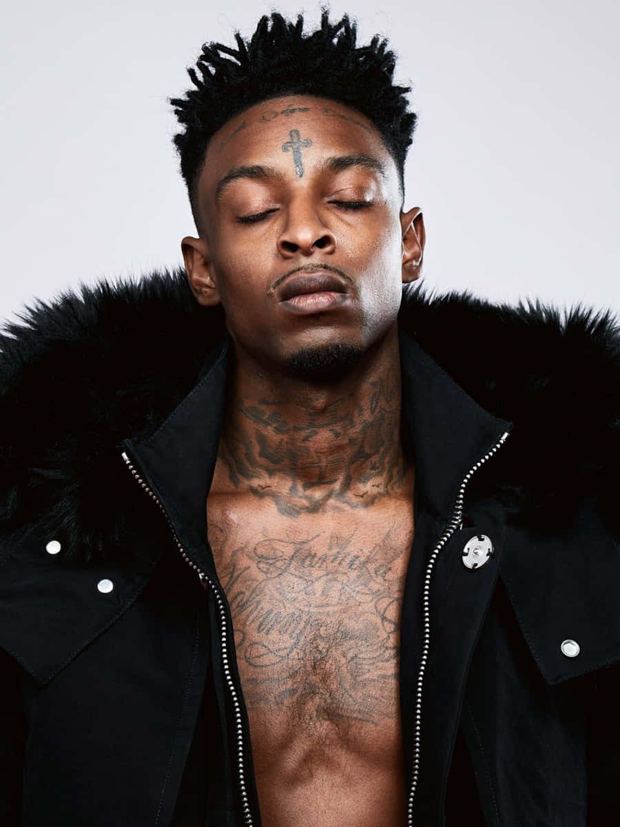 美國饒舌歌手 21 Savage 壁紙 — 一名男子閉上眼睛，胸前和額頭上有紋身，穿著黑色毛領外套，背景簡潔。