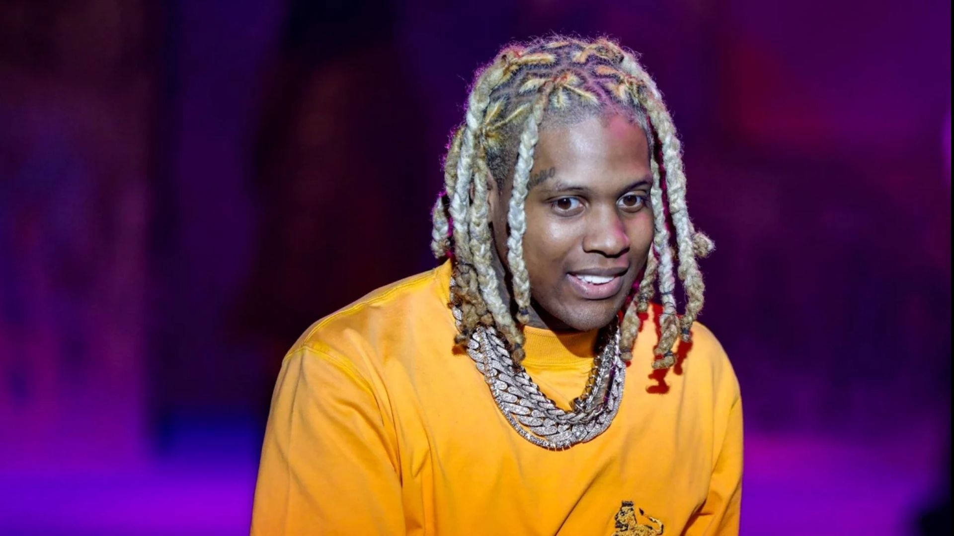 美國饒舌歌手 Lil Durk 壁紙 — 一名擁有彩色辮子頭髮和層疊項鍊的男子，穿著亮橙色襯衫，在紫色燈光背景下微笑。