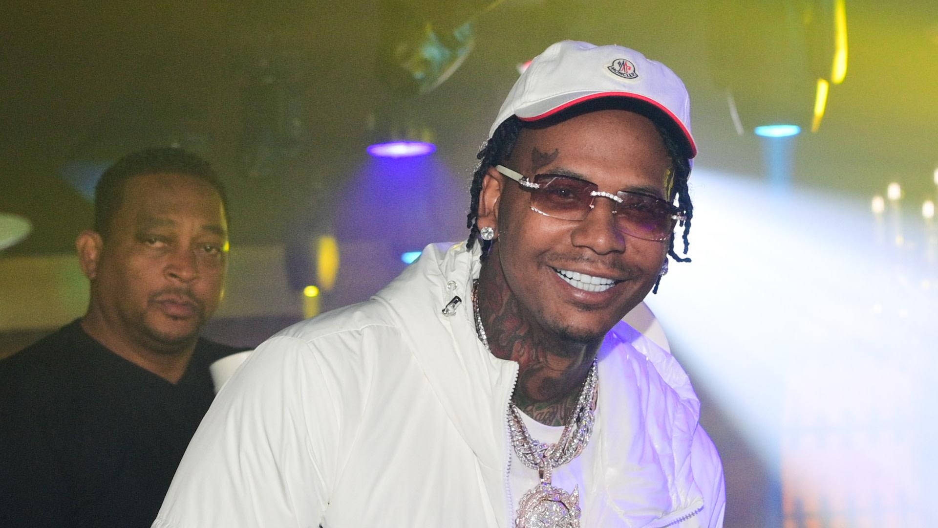美國饒舌歌手 Moneybagg Yo 壁紙 — 一名戴白色帽子和穿白色外套的微笑男子在彩色舞台燈光下擺姿勢，身後有另一名男子。