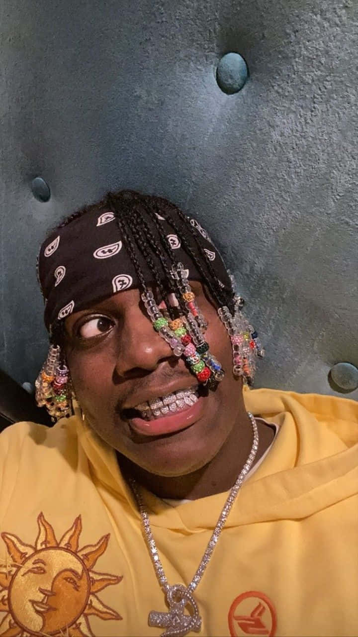 美國饒舌歌手、創作人Lil Yachty 壁紙 — 一個留著辮子、飾有彩色珠子和鑽石牙套的男人，穿著帶有太陽圖案的黃色連帽衫微笑。