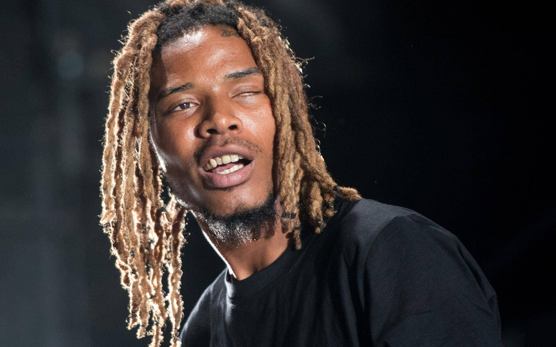 美國饒舌歌手Fetty Wap 壁紙 — 一個長著金色長髮辮的男人在黑暗背景下熱情歌唱的特寫。
