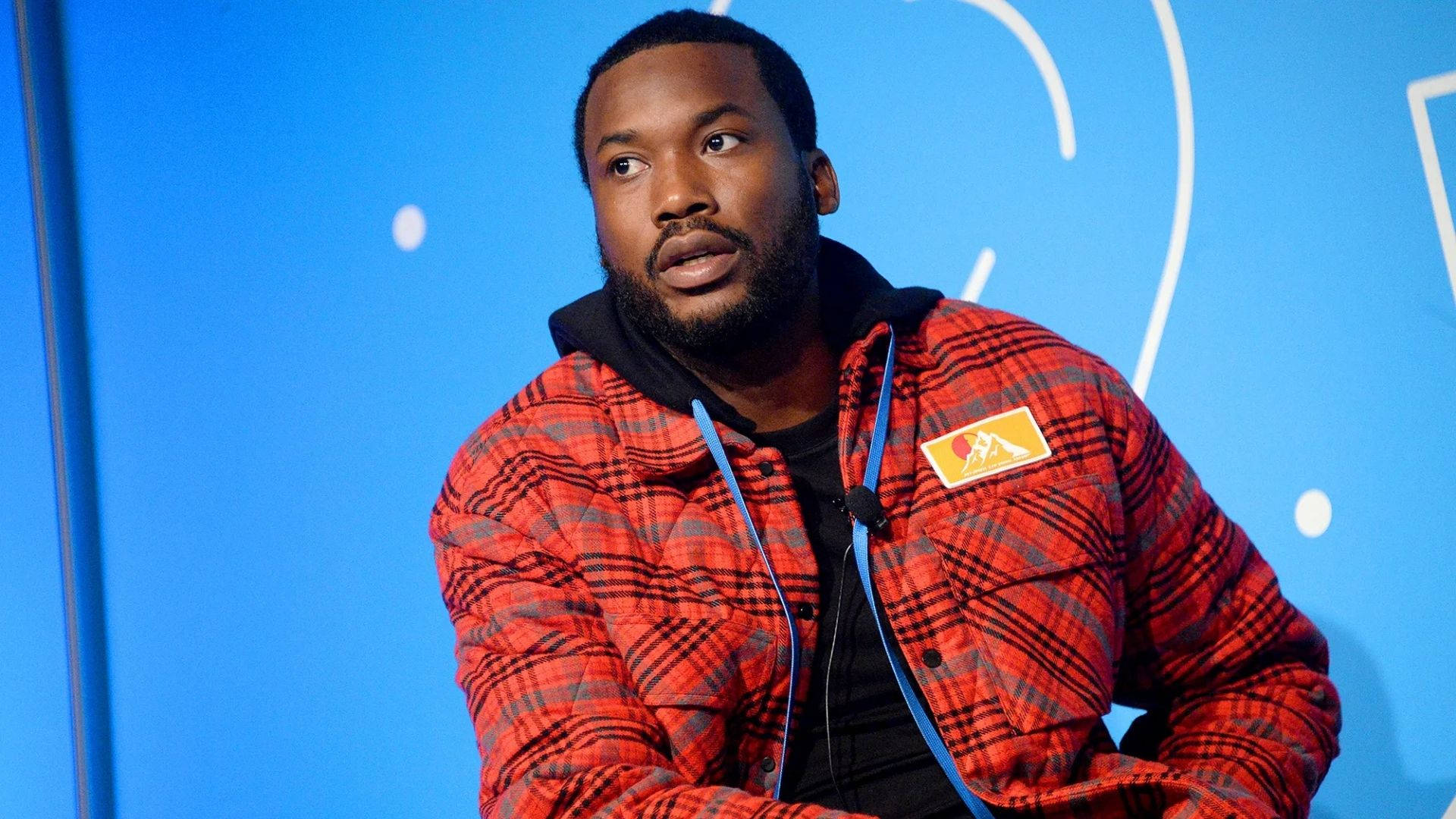 美國饒舌歌手Meek Mill 壁紙 — 一位留鬍鬚的男子穿著紅色格紋夾克，背景為藍色帶白色抽象線條。