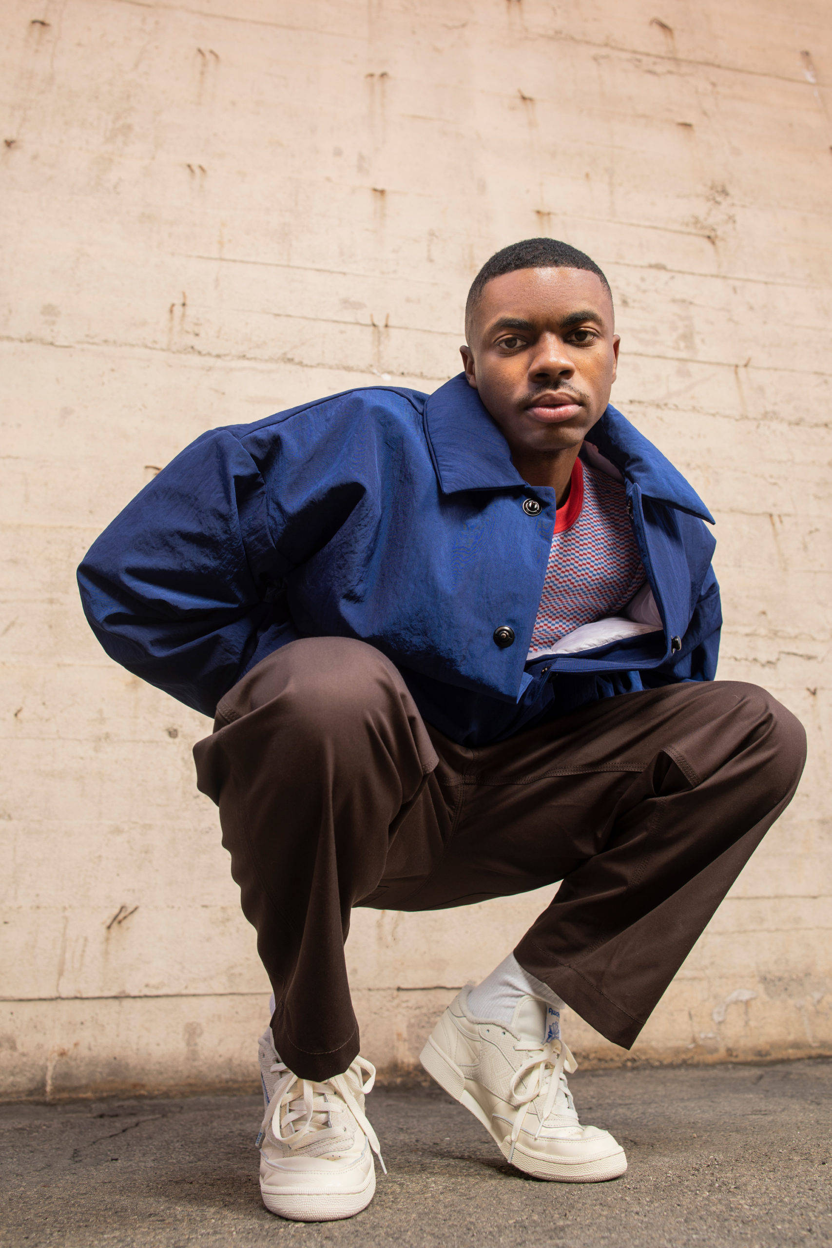 美國饒舌歌手Vince Staples 壁紙 — 一位身穿藍色外套和白色運動鞋的年輕男子自信地蹲在風化的混凝土牆前。