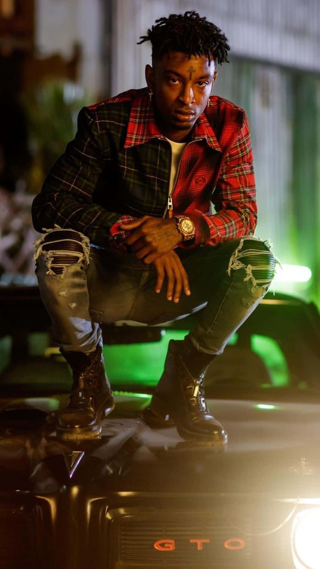 美國饒舌歌手兼作曲人 21 Savage 壁紙 — 一名身穿紅色格仔外套和破洞牛仔褲的年輕男子，在霓虹綠燈光下自信地蹲在一輛黑色GTO汽車上。