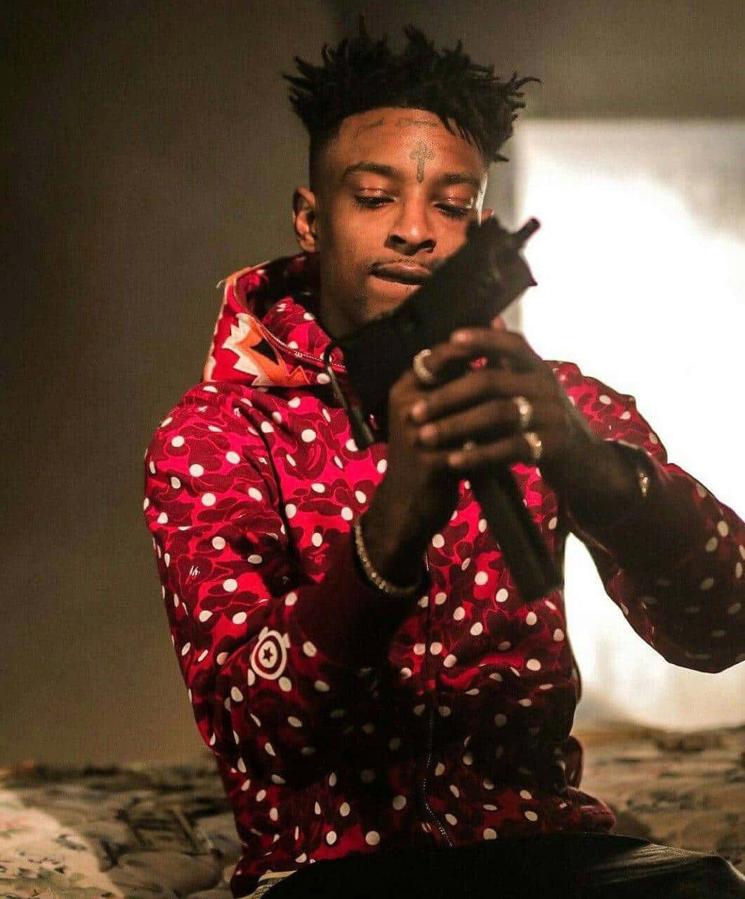 美國饒舌歌手兼唱片製作人 21 Savage 壁紙 — 一名身穿紅色圖案連帽衫的男子在戲劇性燈光下持槍，表情嚴肅。