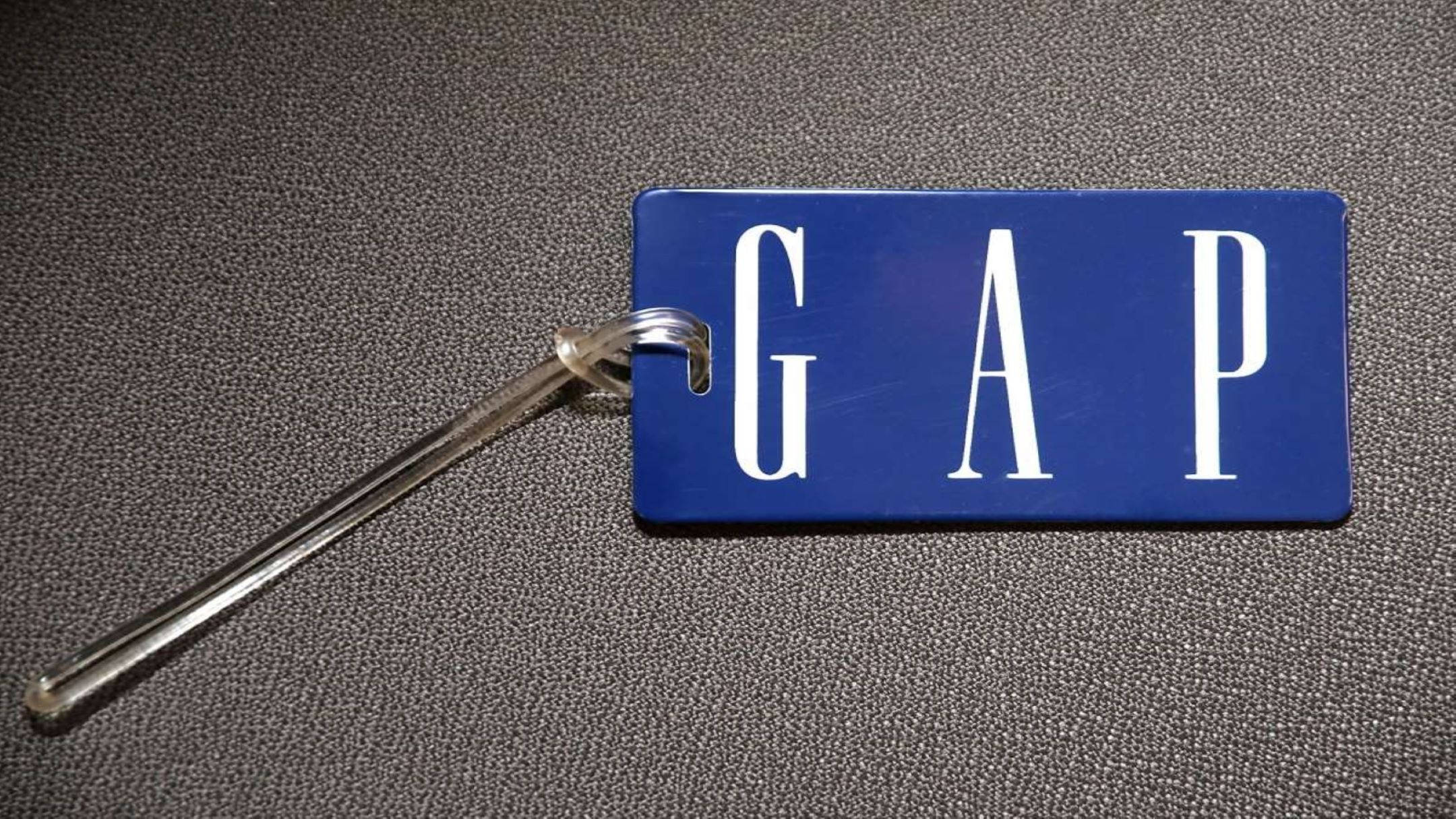 美學 Gap 藍色吊牌 壁紙 — 一個藍色Gap行李牌，上面有白色字體，放置在帶有紋理的灰色表面上，並由透明塑料環固定。