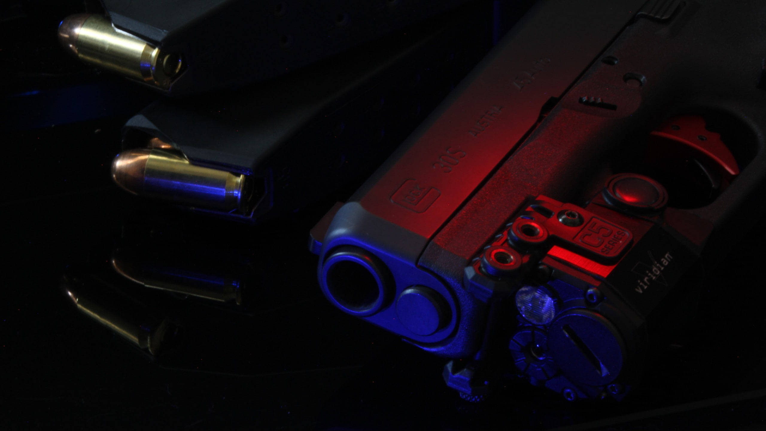美學 Glock 與子彈 壁紙 — 手槍和子彈在黑色反光表面上，在戲劇性的紅色和藍色燈光下閃閃發光。