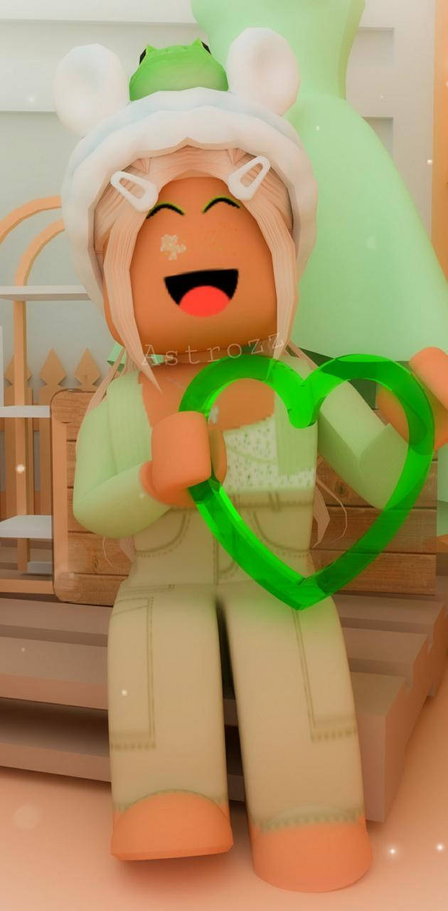 美學 Roblox 女孩配綠心 壁紙 — 一個戴著兔耳和青蛙帽的開心Roblox角色在舒適的房間裡拿著一個發光的綠色心形。