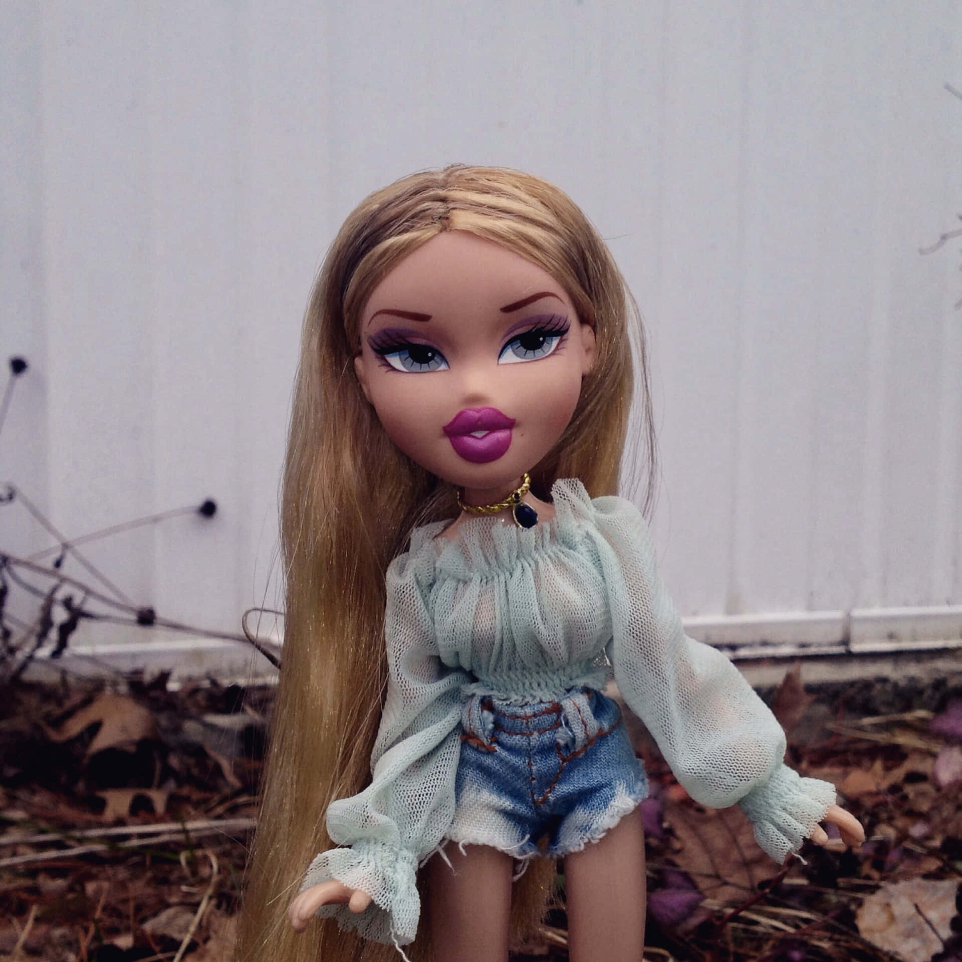 美學Bratz玩偶Yasmin 壁紙 — 一個擁有金色長髮和紫色嘴唇的 Bratz 公仔，穿著淺藍色網眼上衣和牛仔短褲，背景是白色籬笆和落葉。