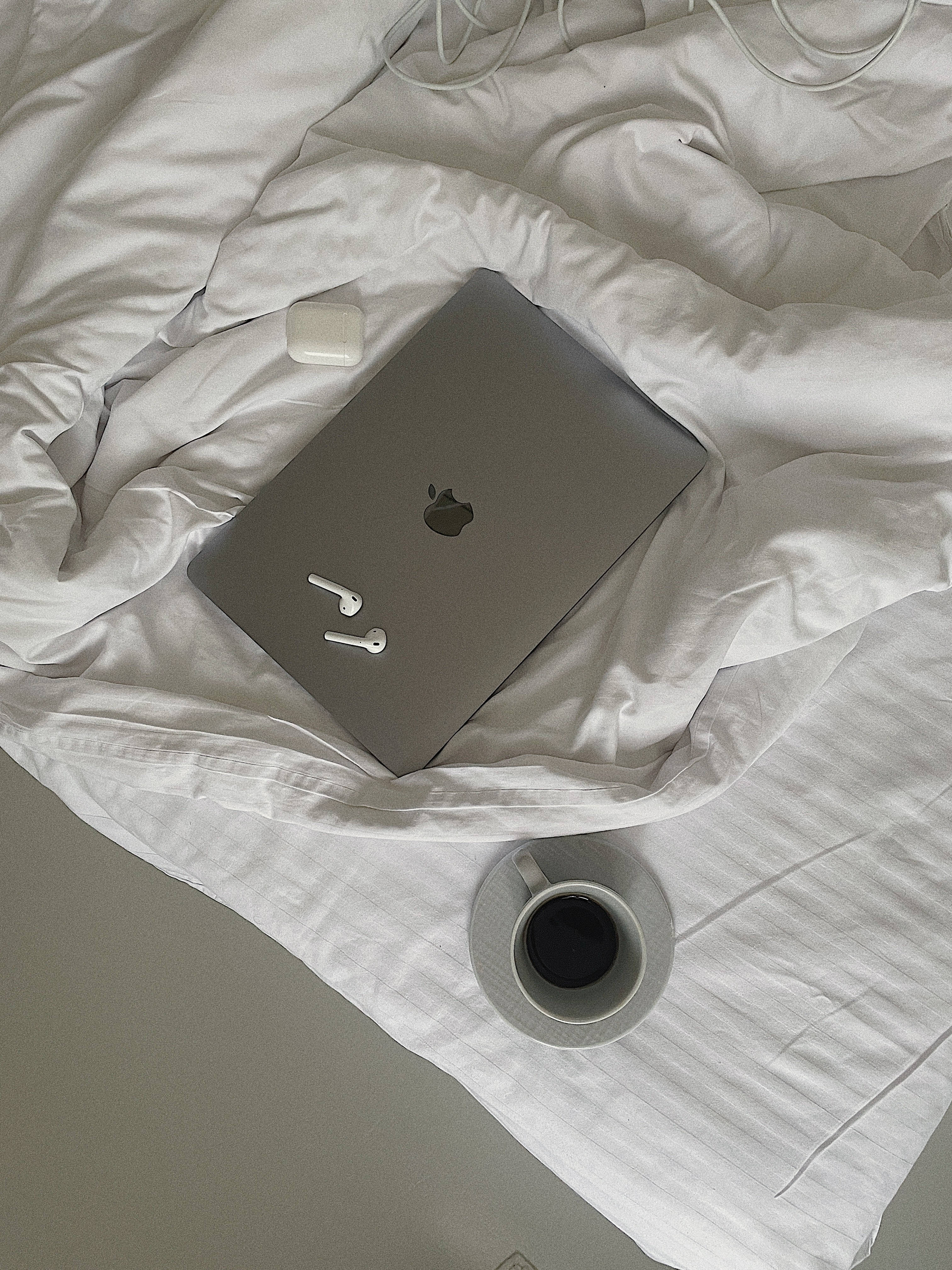 美學Tumblr筆電MacBook咖啡 壁紙 — 一部銀色手提電腦和白色耳塞放在皺巴巴的白色床單上，旁邊有一杯咖啡。