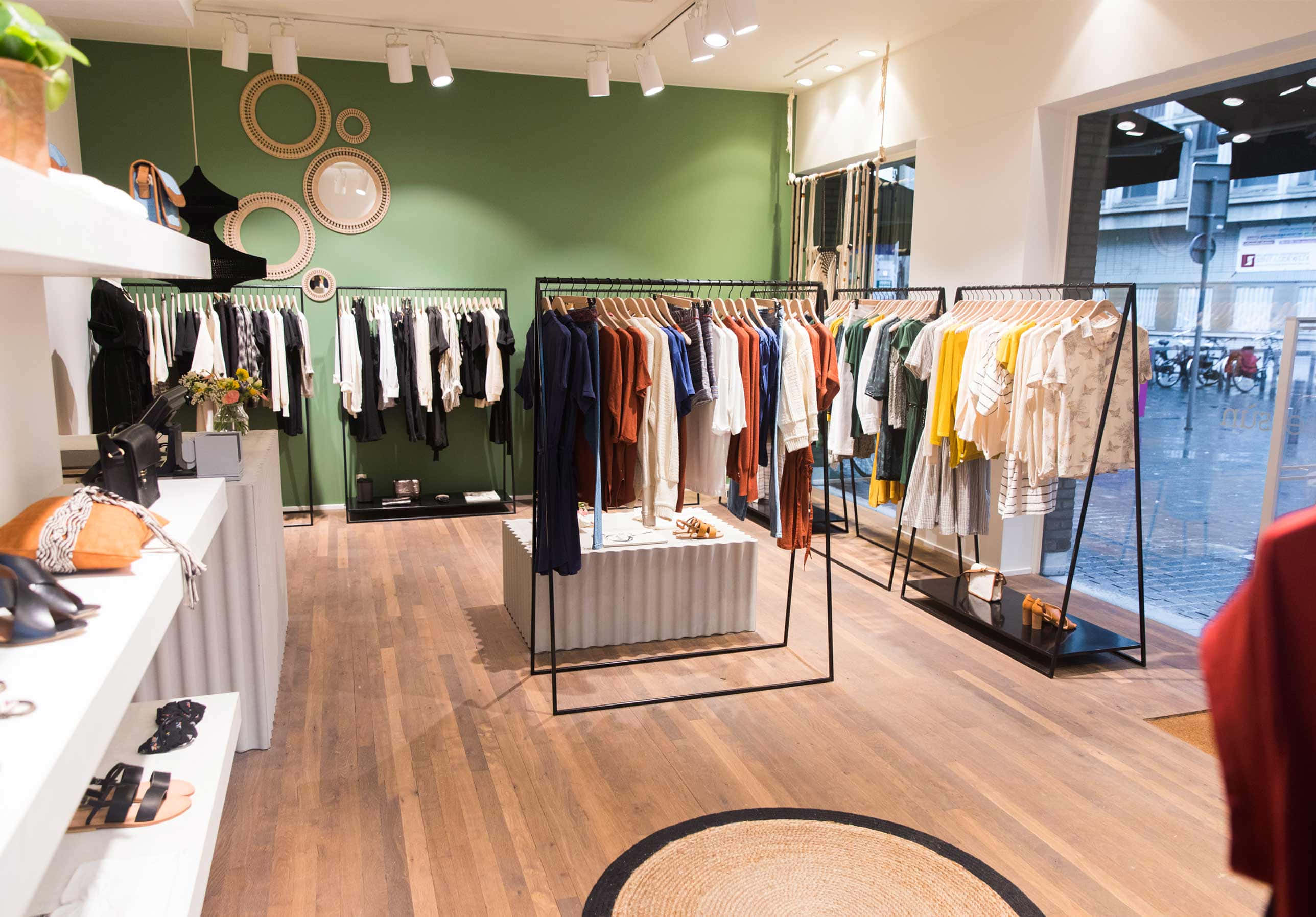 美學服裝店圖片 壁紙 — 一間現代精品店，舖有木地板、綠色牆壁，在明亮的吊燈下展示色彩繽紛的衣物架。