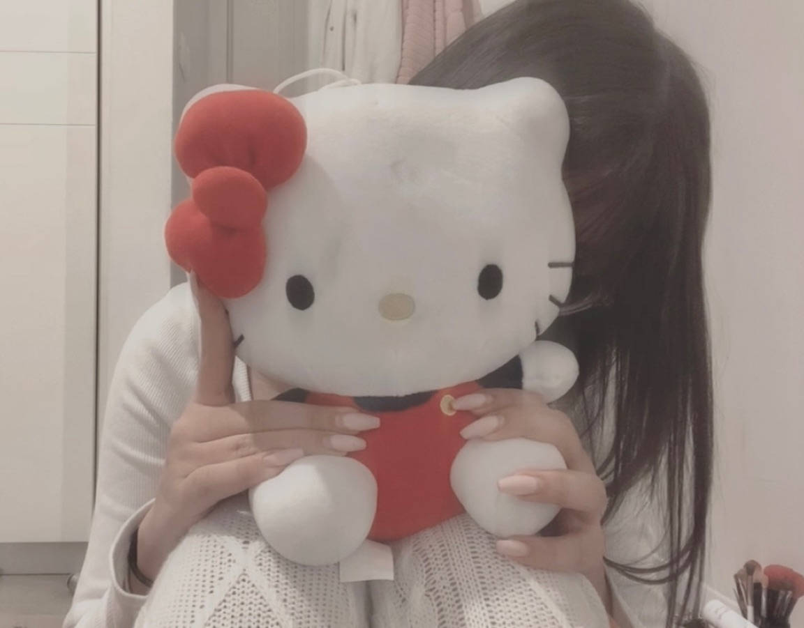 美學玩偶Hello Kitty頭像 壁紙 — 一位長黑髮的人士在室內擁抱一個戴紅色蝴蝶結和穿紅色連衣裙的大型白色Hello Kitty毛公仔。