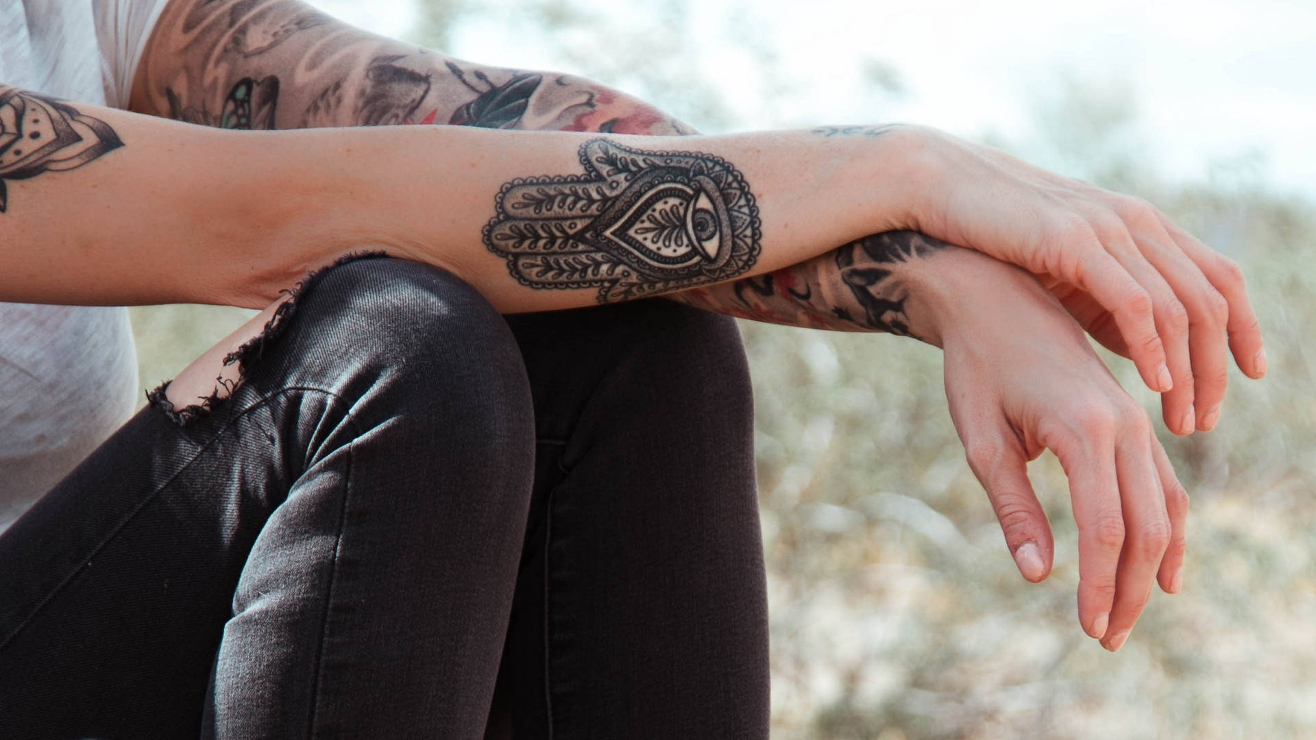 美學風格的「Hamsa」手印紋身 壁紙 — 特寫紋身手臂放在膝蓋上，帶有複雜的Hamsa手設計，中心有一隻眼睛。