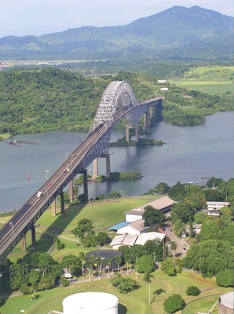 美洲橋橫跨巴拿馬運河的壯麗景色 壁紙 — 晴朗藍天下，一座鋼拱橋橫跨寬闊河流，背景是鬱鬱蔥蔥的綠色山丘和山脈的鳥瞰圖。