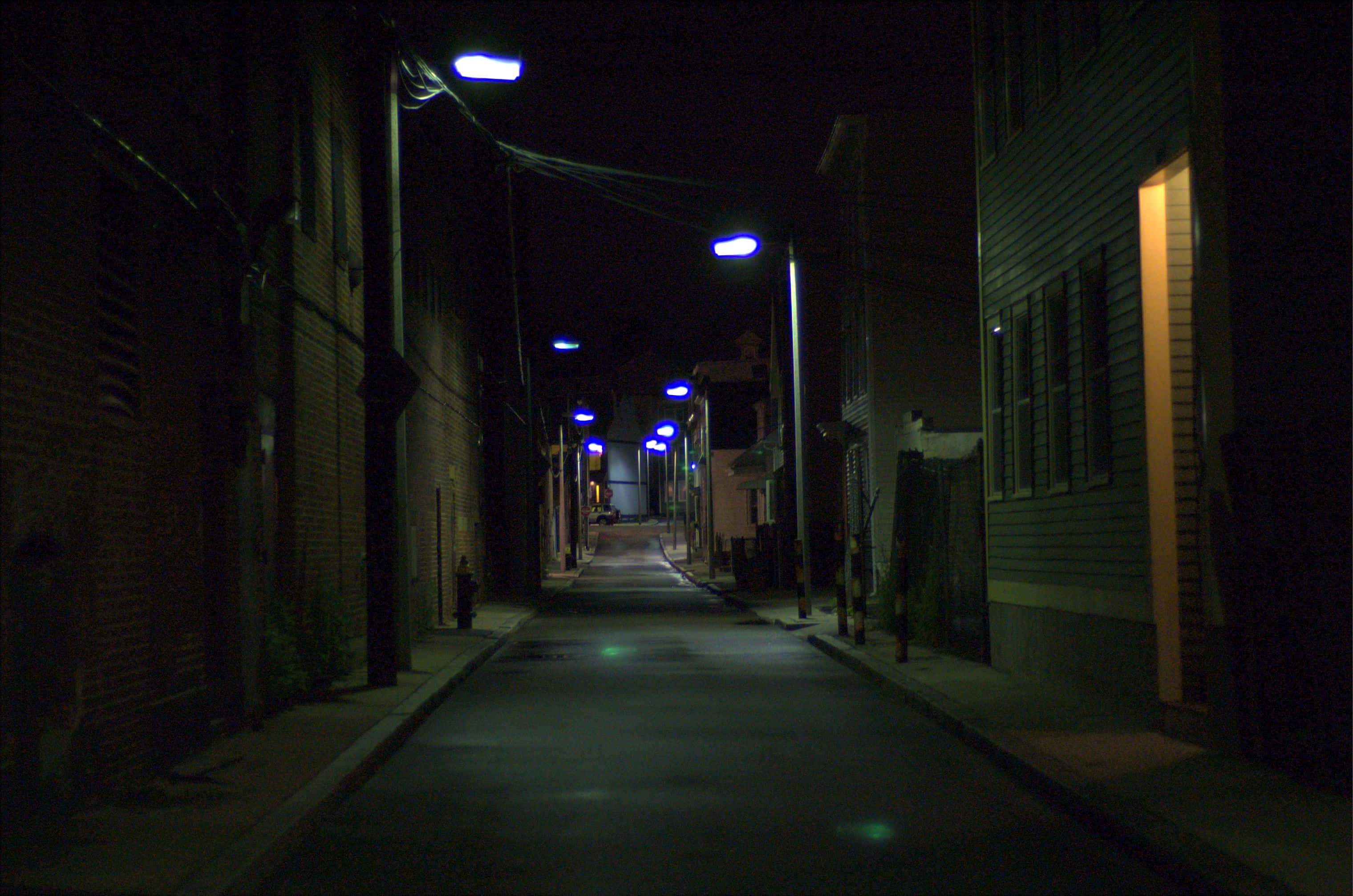 美麗孤寂的巷道 壁紙 — 夜晚一條狹窄的城市小巷，被藍色街燈照亮，在磚木建築之間投下長長的影子。