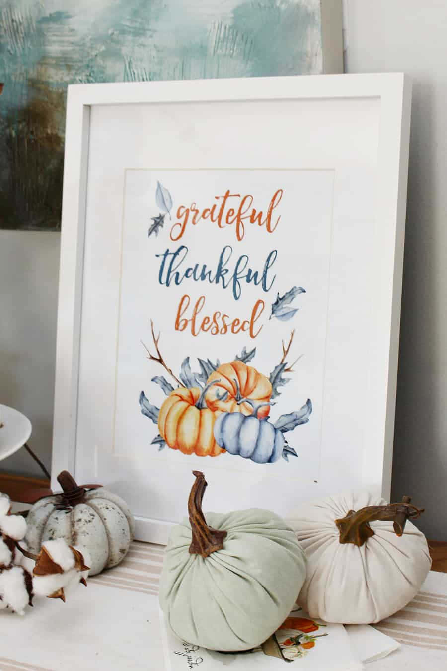 美麗感恩節相框圖 壁紙 — 一個帶有 'grateful thankful blessed' 文字和南瓜的裱框水彩印刷品，旁邊有布料南瓜和棉花，放在條紋桌上。