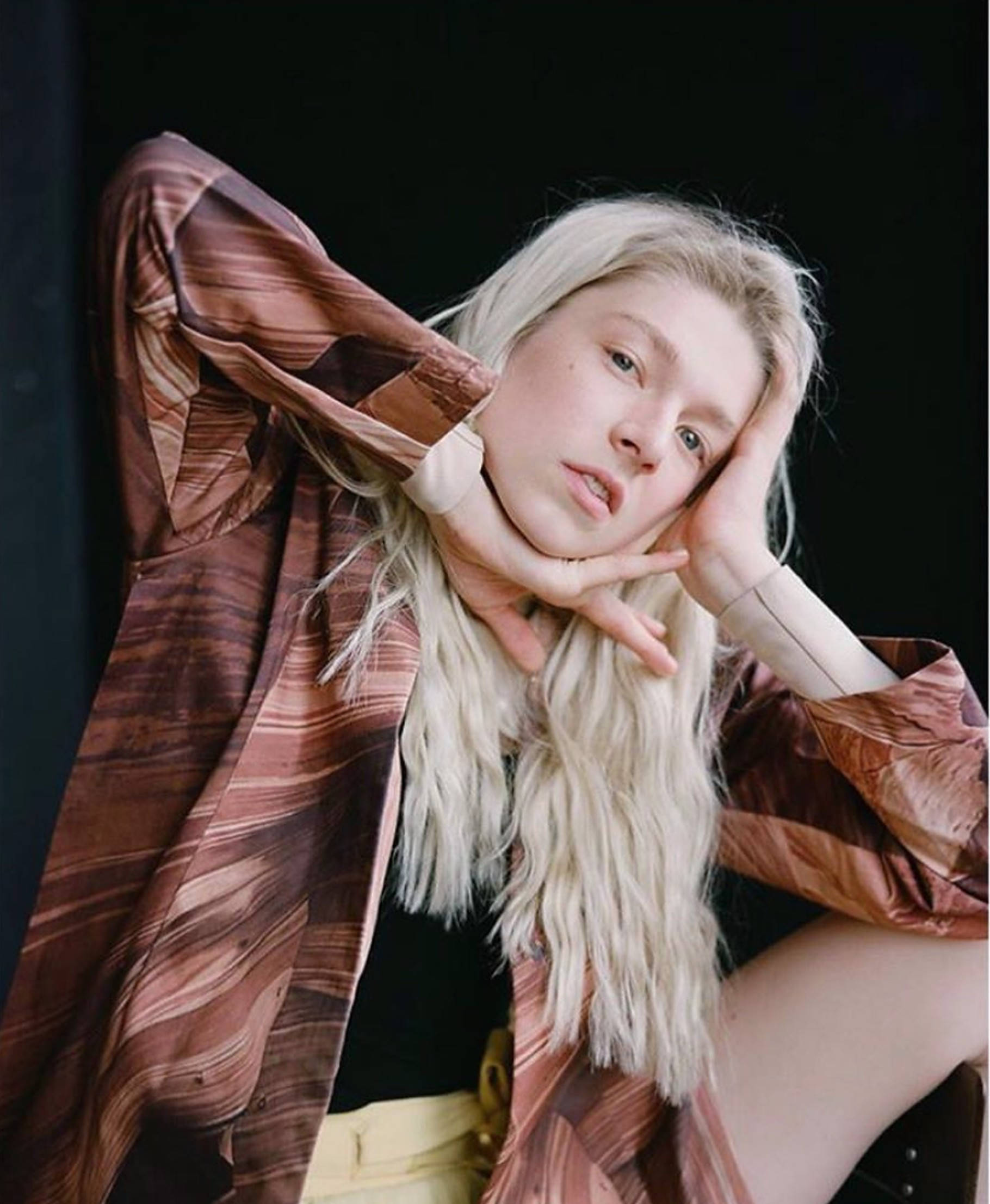 美麗的 Hunter Schafer 壁紙 — 一位年輕女性，一頭鉑金色頭髮，在純黑背景前將下巴擱在手上，身穿飄逸的棕色圖案襯衫。