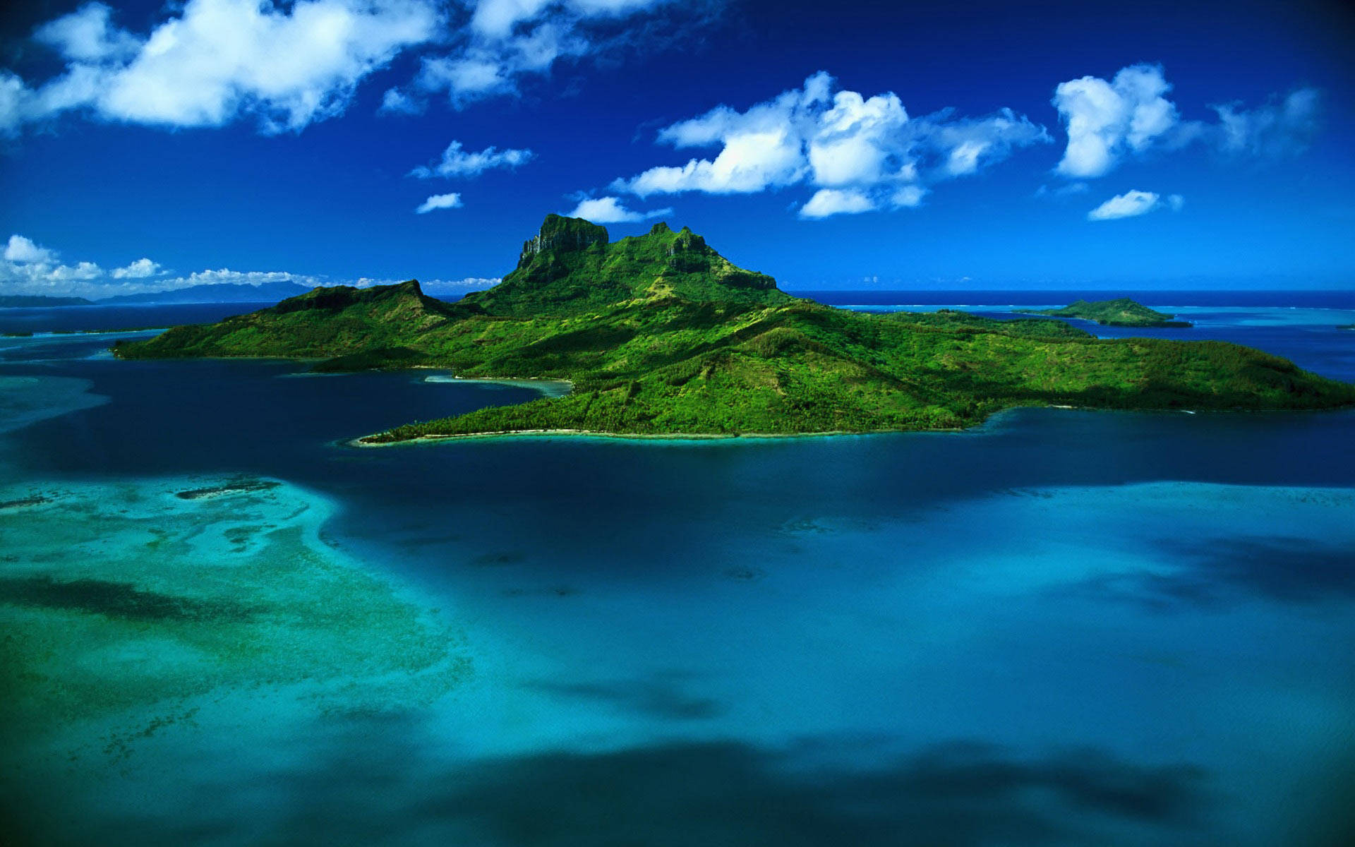 美麗的加勒比海島風景 壁紙 — 翠綠島嶼的鳥瞰圖，島上有火山峰，周圍環繞著充滿活力的綠松石色潟湖和深藍色海洋，天空明亮，有蓬鬆的白雲。