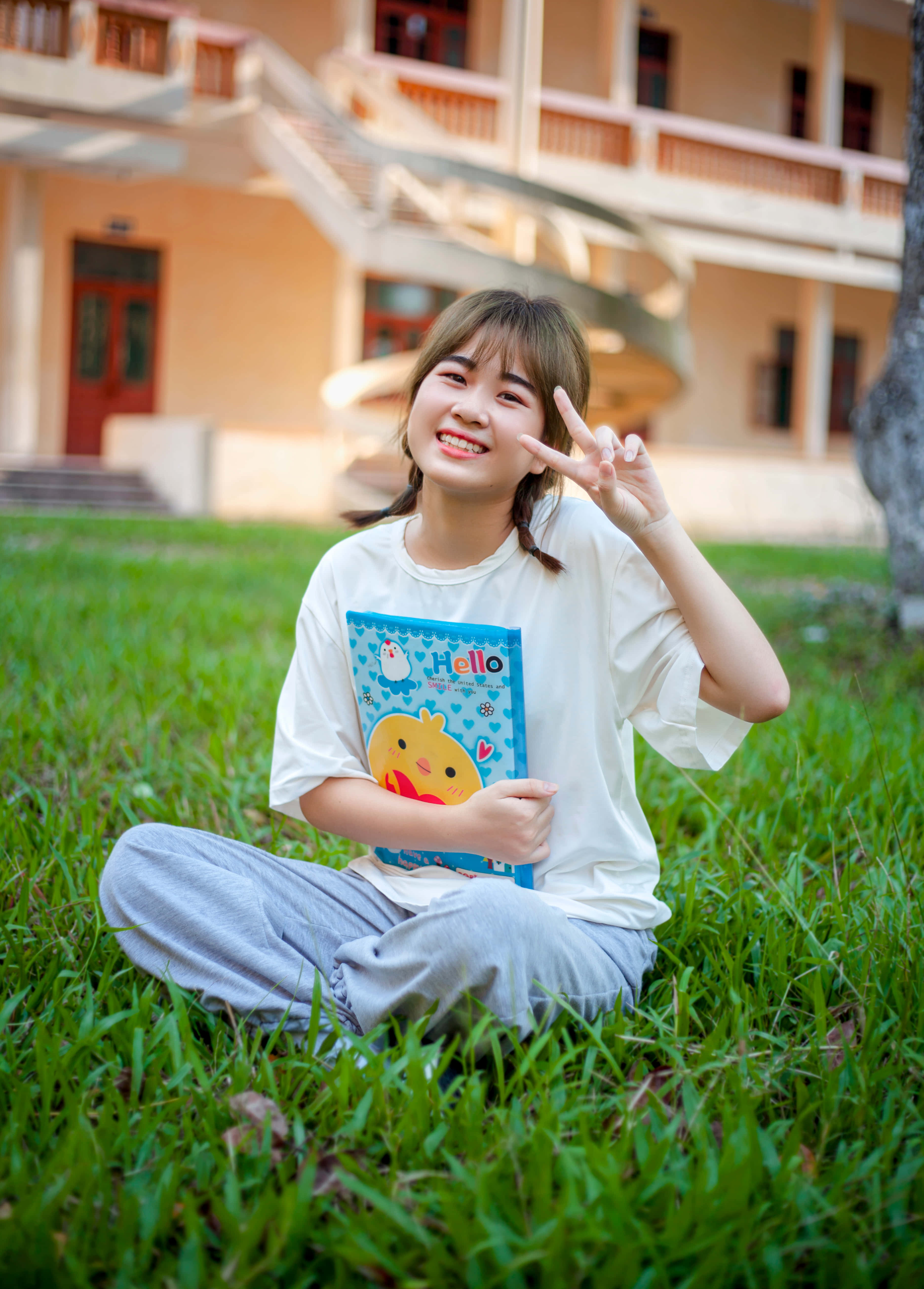 美麗的年輕女孩，快樂無憂 壁紙 — 一位微笑的年輕女性盤腿坐在米色建築前充滿活力的綠色草地上，手持彩色筆記本並做出和平手勢。