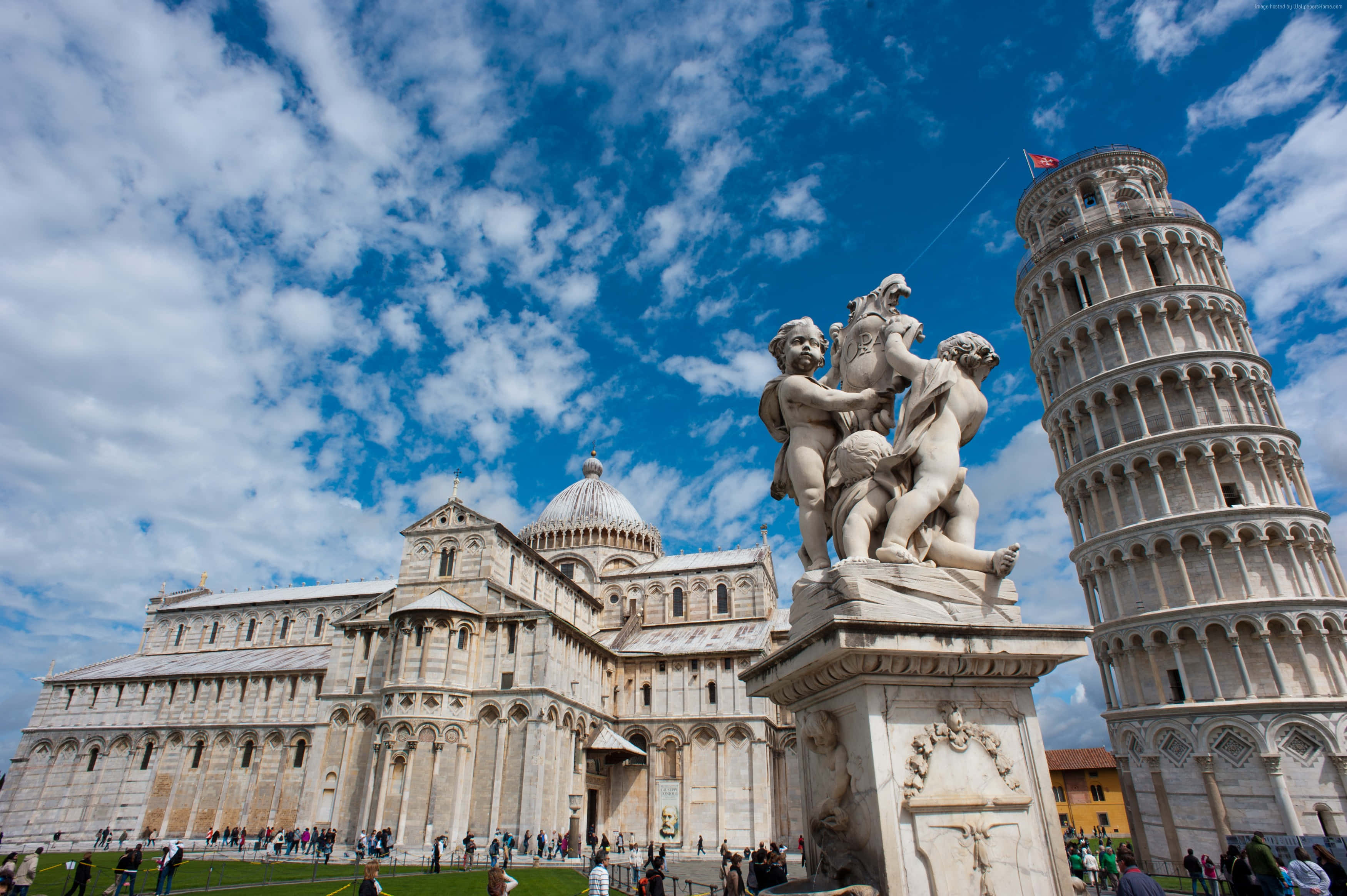 美麗的日落 壁紙 — 比薩斜塔矗立在比薩大教堂旁邊，在明亮的藍天下，有散落的雲朵和前景的大理石雕像。
