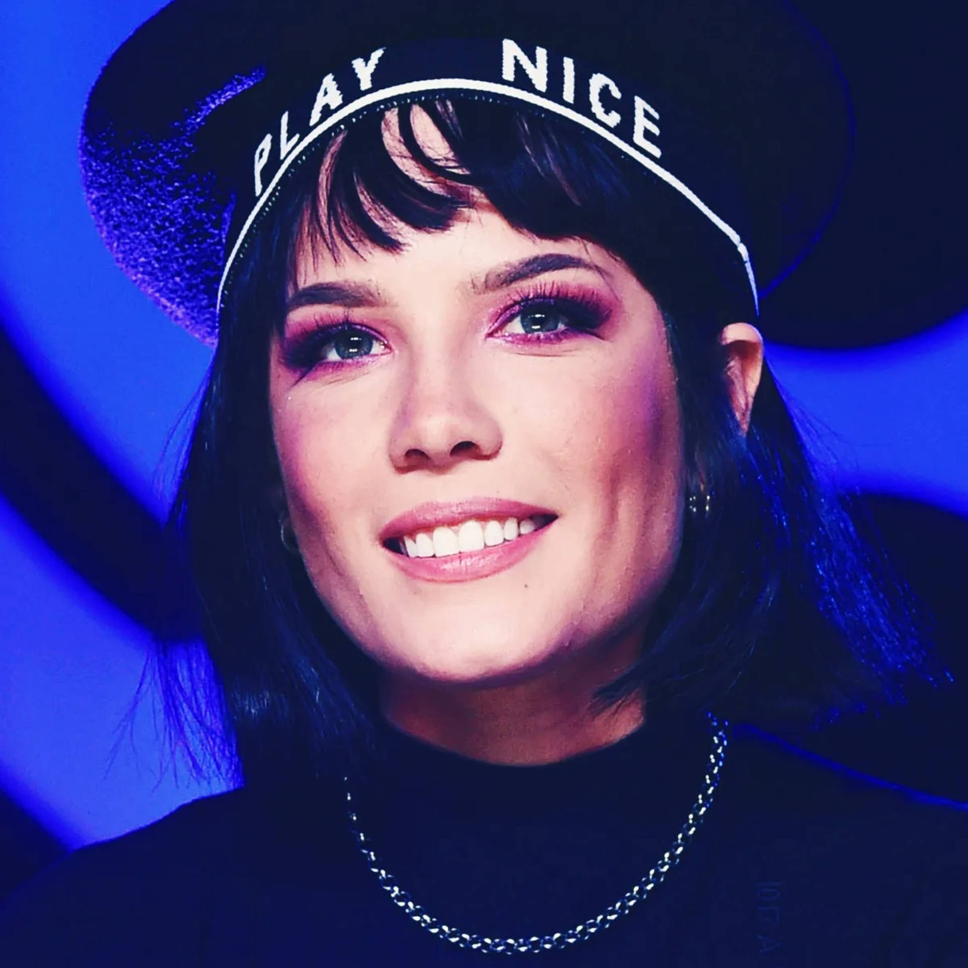 美麗的歌手 Halsey 壁紙 — 一位深色頭髮、妝容大膽的女性，戴著一頂帶有白色「PLAY NICE」字樣的黑色帽子，在充滿活力的藍色背景下溫暖地微笑。