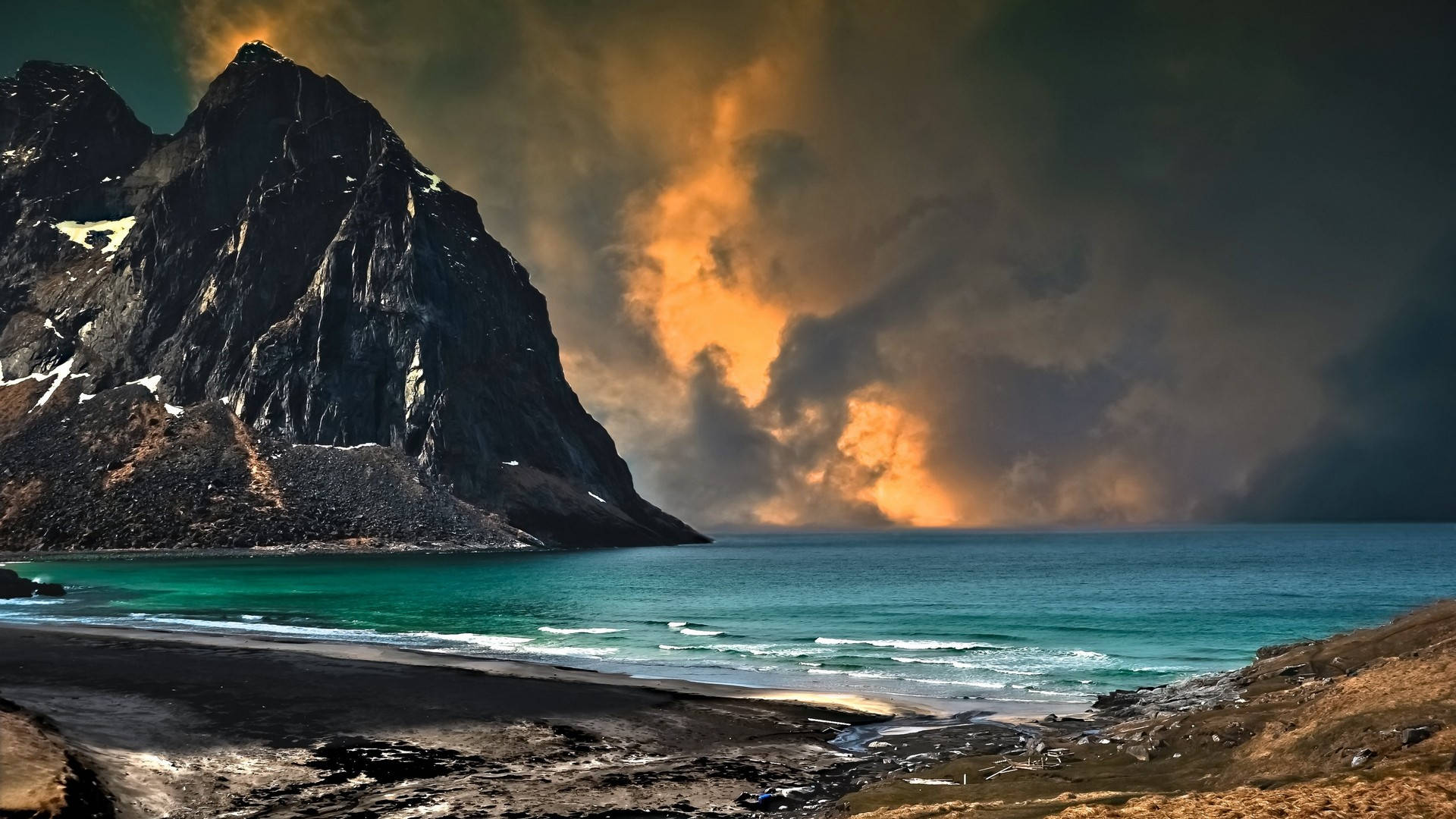 美麗的海濱天空 壁紙 — 一座壯麗的山峰聳立在黑色沙灘之上，金色夕陽穿透烏雲，照耀在碧綠的海水上。
